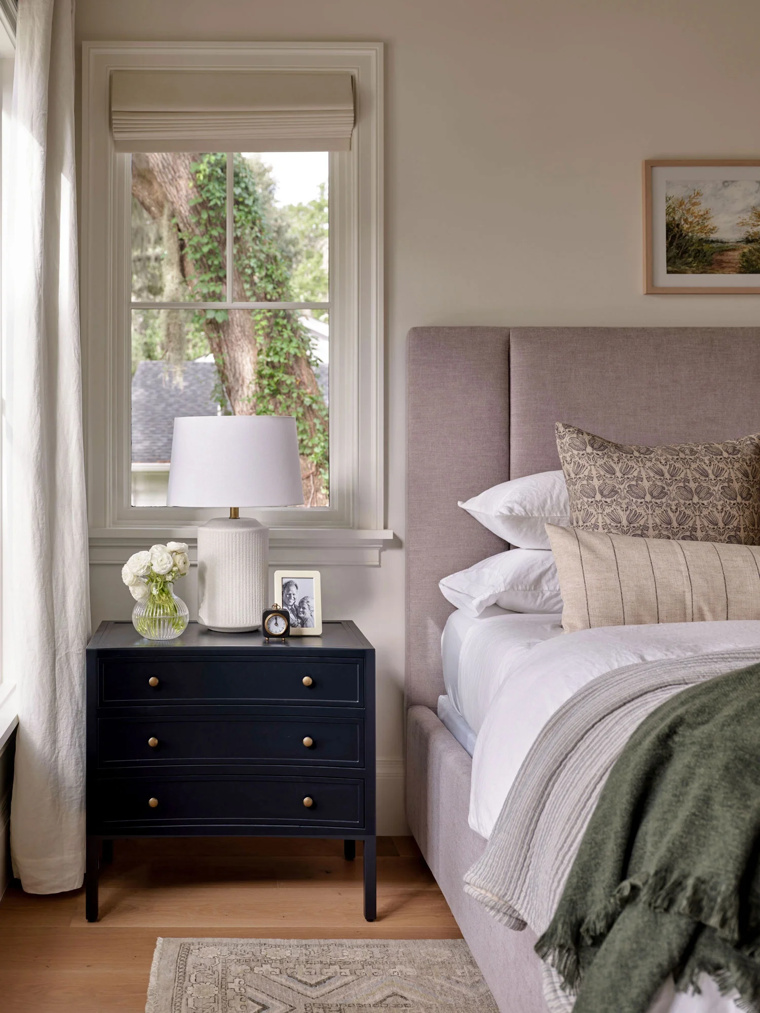 A bedroom corner with a beige upholstered bed, patterned pillows, a navy nightstand with a white lamp, framed photo, and a clock, a window with view of a large tree outside, and a landscape painting on the wall.