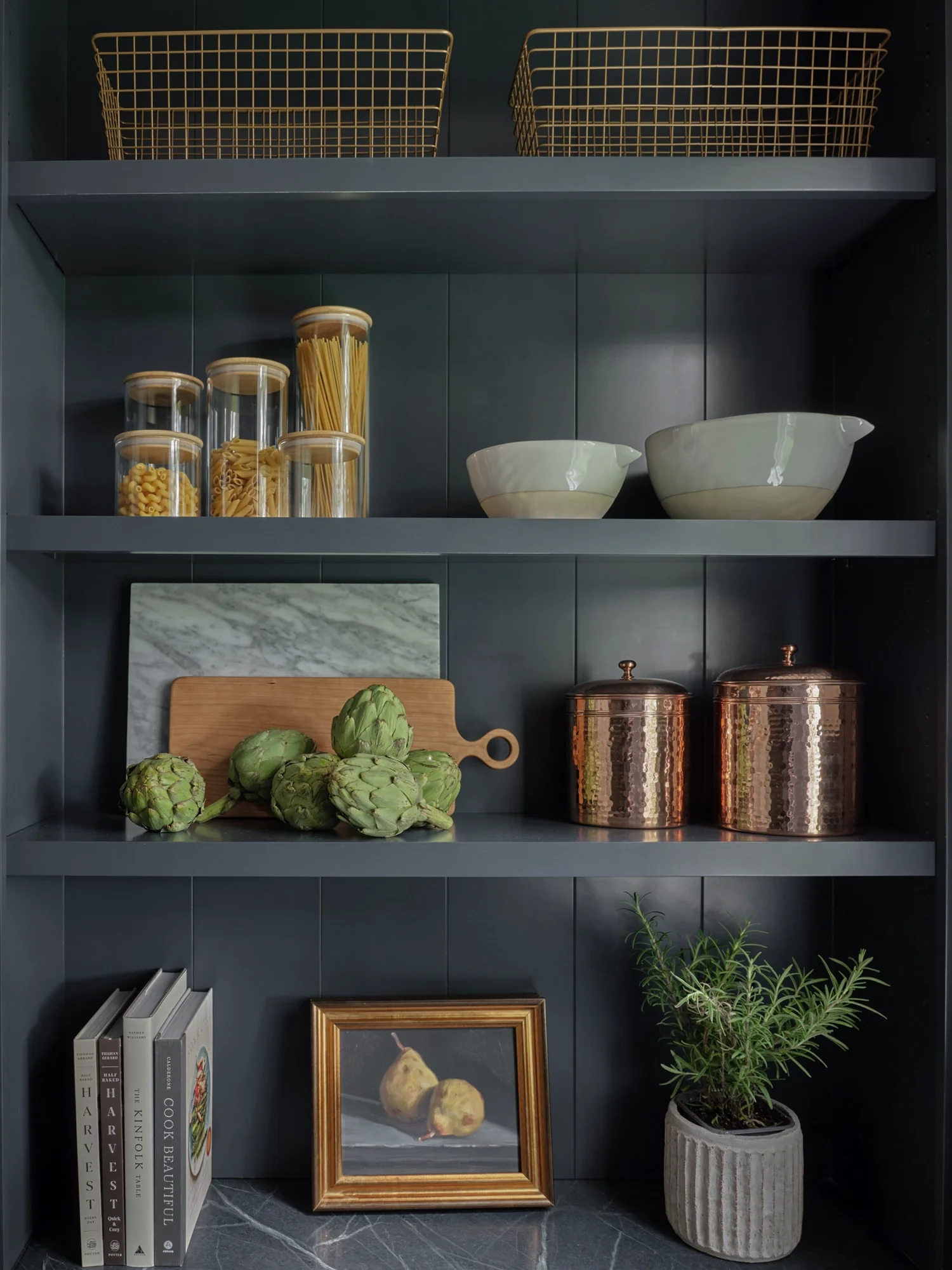 A kitchen shelf with two wire baskets on top, glass jars of pasta on the middle shelf, ceramic bowls, artichokes on a cutting board, a framed painting of pears, books, and a potted plant.