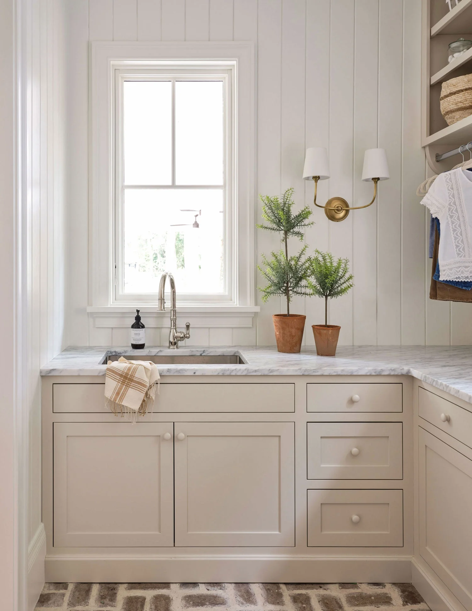 A kitchen with white cabinets, a marble countertop, a silver sink with a faucet, a decorative towel, and two potted plants. There is a window above the sink with white trim, and a beige and brown patterned rug on a hardwood floor.