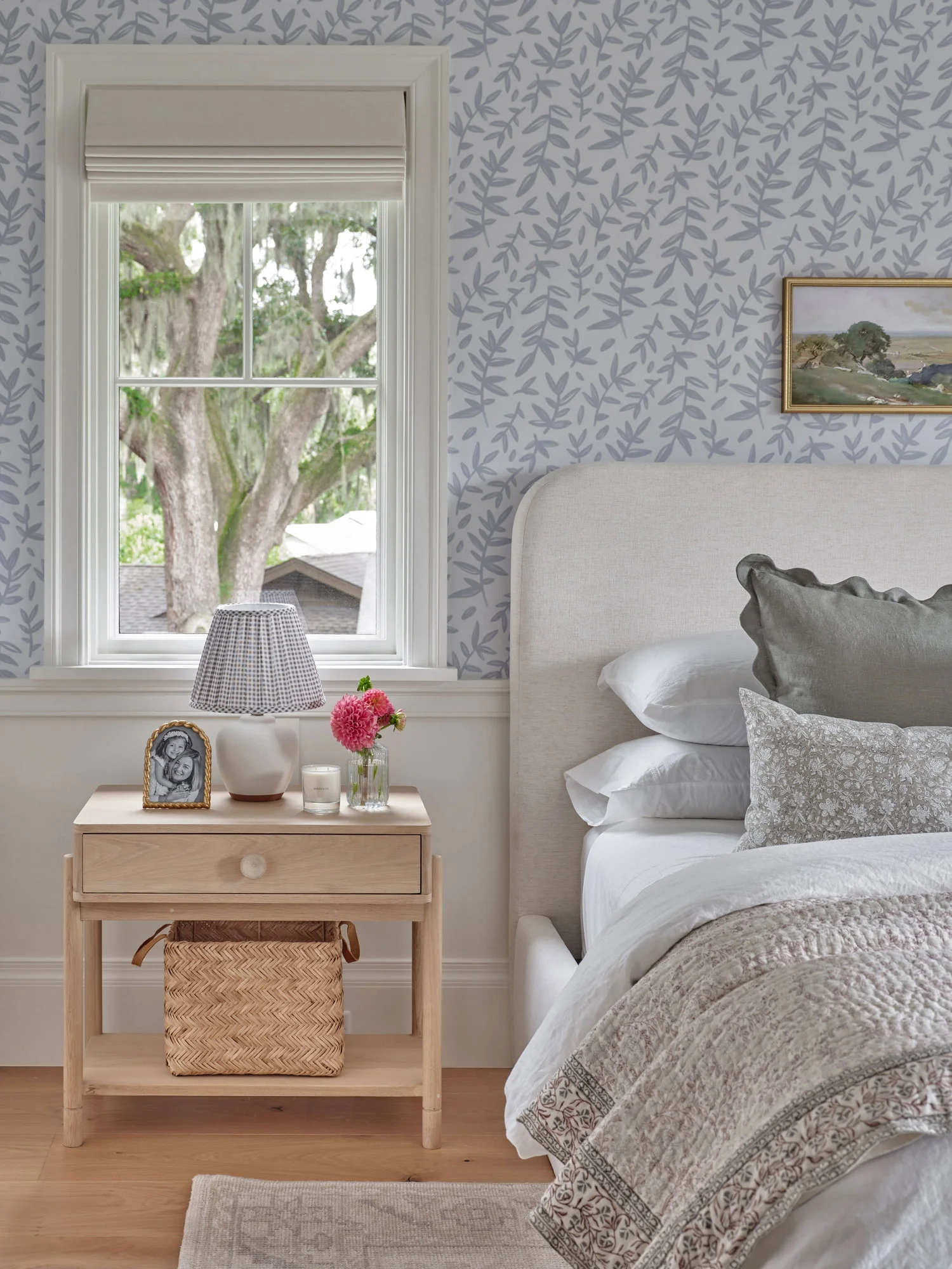 A bedroom with a window showing large trees outside. A nightstand with a lamp, a framed photograph, pink flowers, and candles. Part of a bed with a white headboard and decorative pillows.