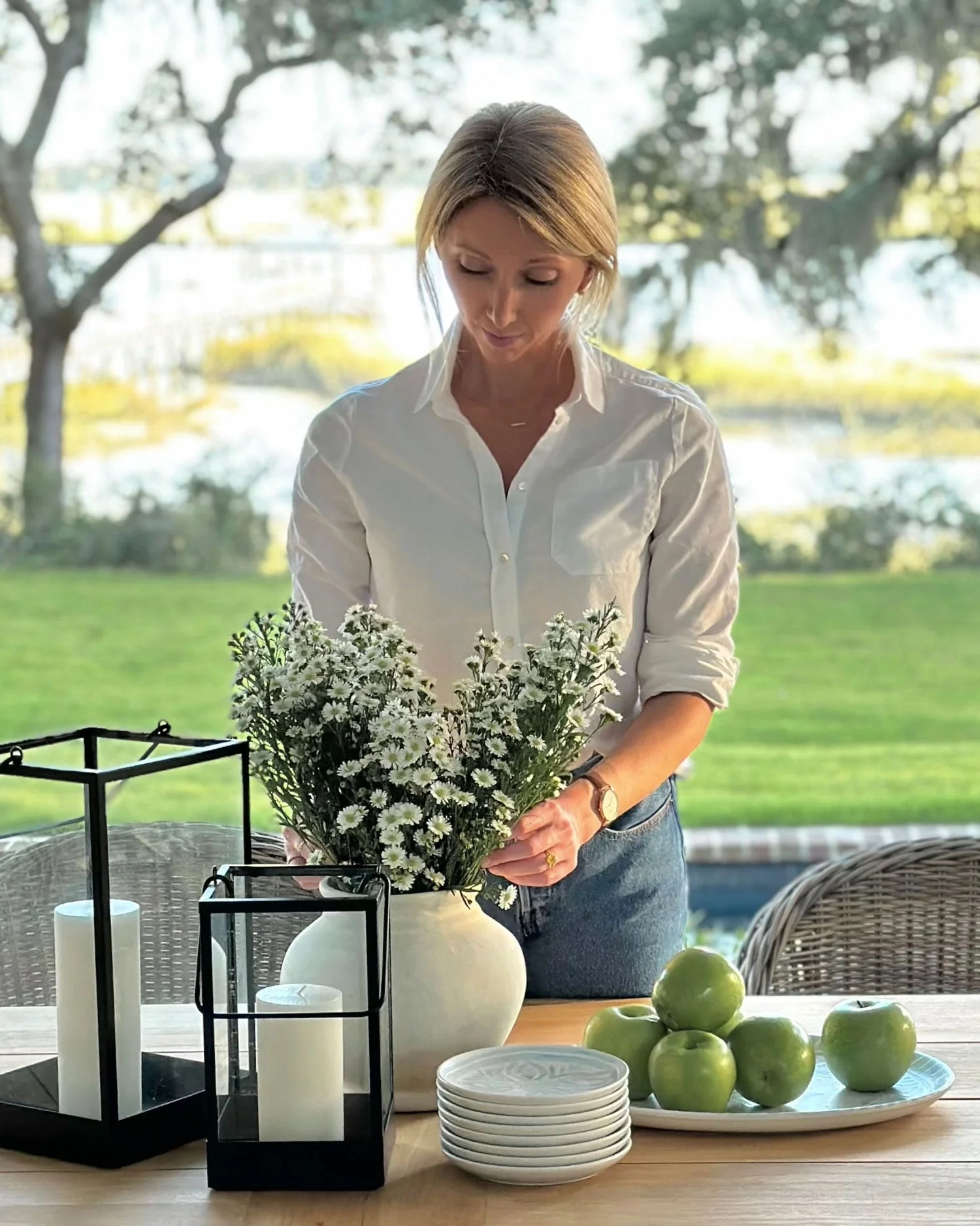 A woman in a white shirt arranging a bouquet of white daisies in a cream-colored vase. The setting appears to be outdoors or near a large window with trees in the background.