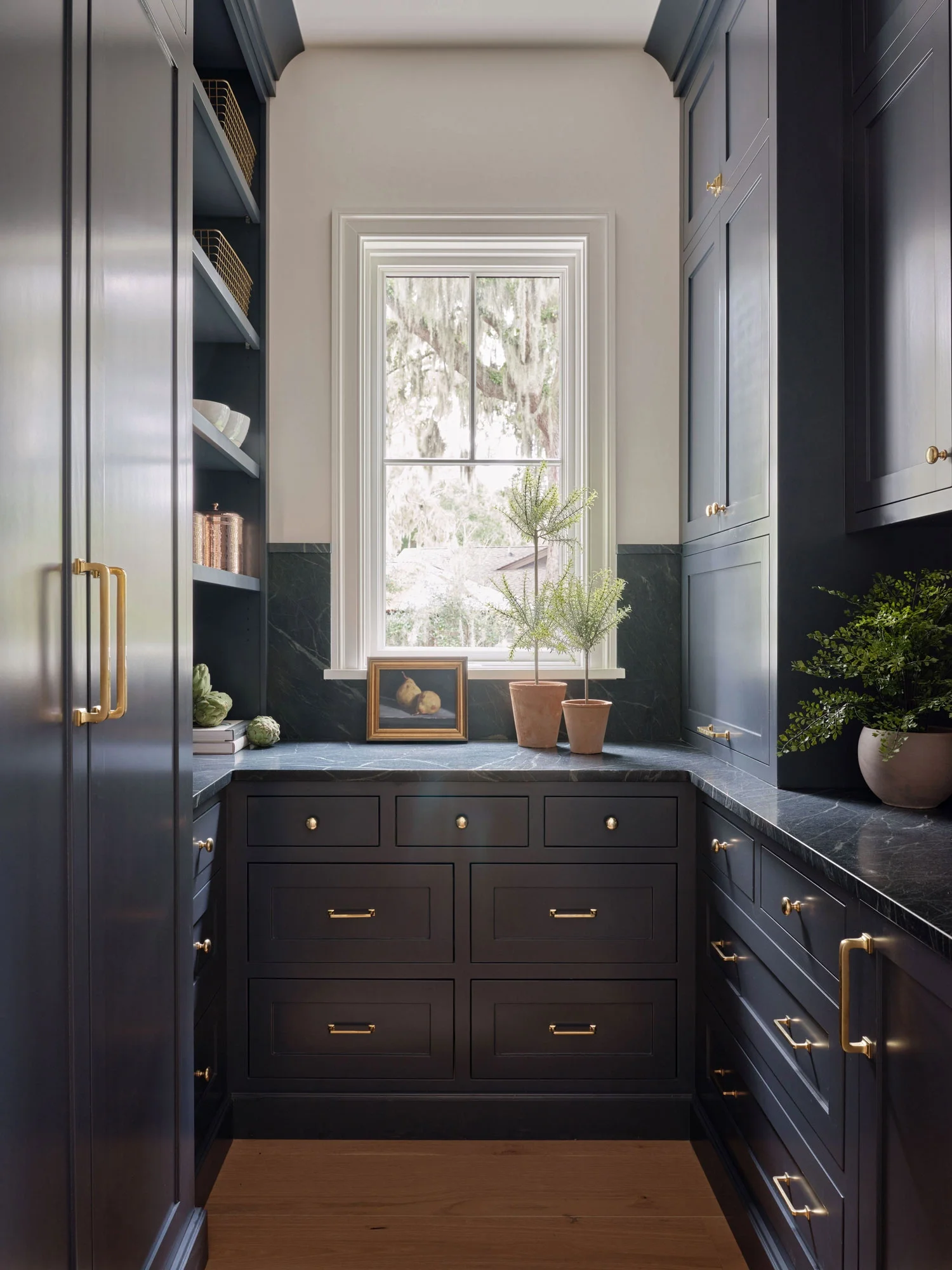 A narrow kitchen workspace with dark blue cabinets, gold hardware, a large window with a view of trees outside, and decorative items including a framed painting, potted plants, and bowls.