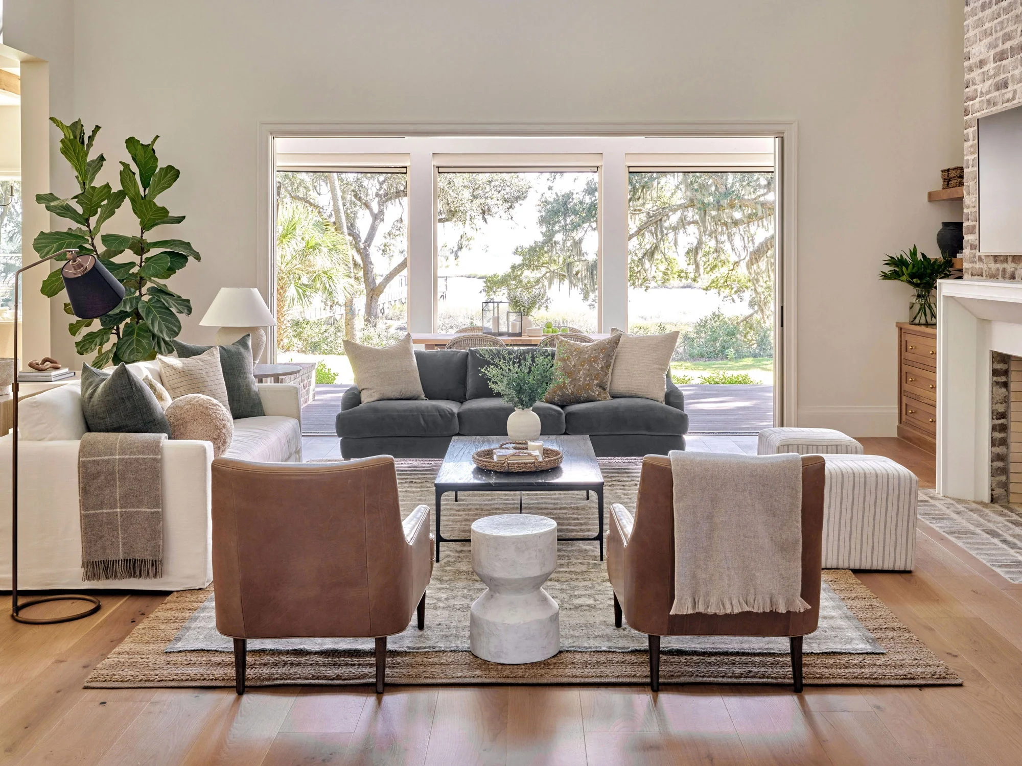 Living room with large windows showing outdoor trees, with seating including a white sofa and brown chairs, a marble side table, hanging plants, and decorative items.