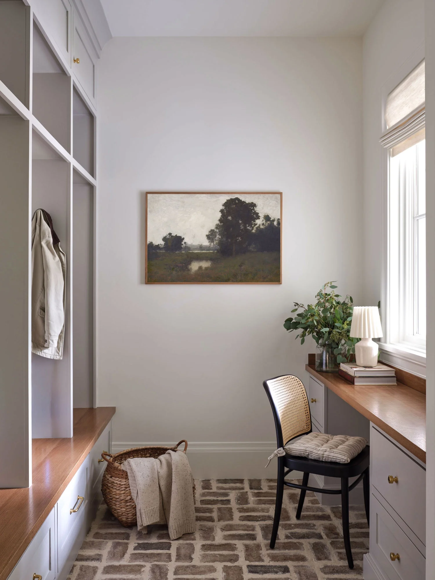 A cozy corner workspace with a wooden desk, black chair with a cushion, a stack of books, a white table lamp, and a large leafy plant near a window with sunlight. A framed landscape painting hangs on the wall, and a wicker basket with clothing is on the floor.