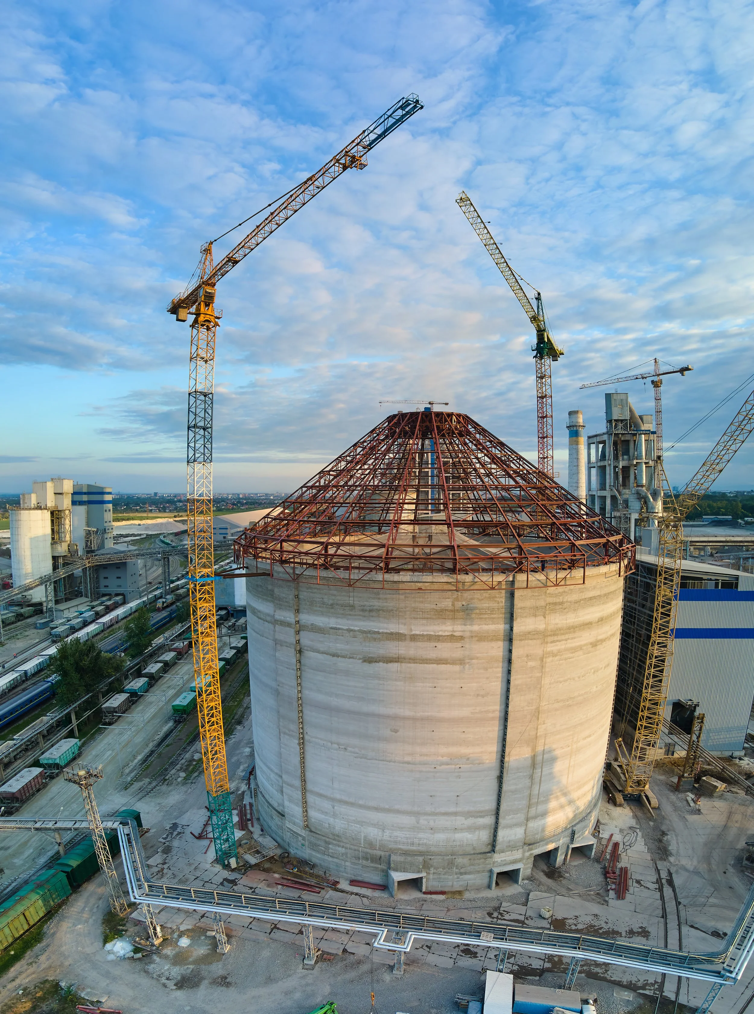 Industrial construction site on a cloudy day. A large, cylindrical concrete structure takes center stage, partially built with a domed metal framework on top.