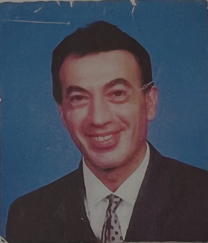 A man in a suit with a white shirt and a patterned tie, smiling against a blue background.