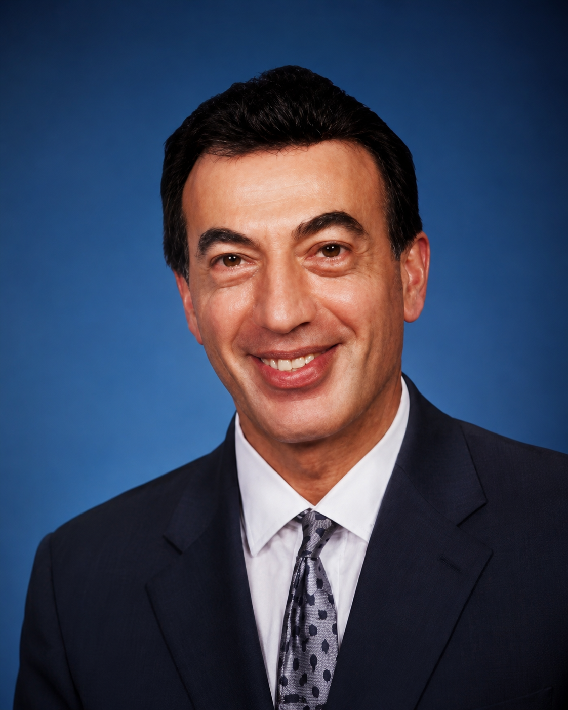 A professional headshot of a smiling man in a dark suit, white shirt, and patterned tie against a blue background.