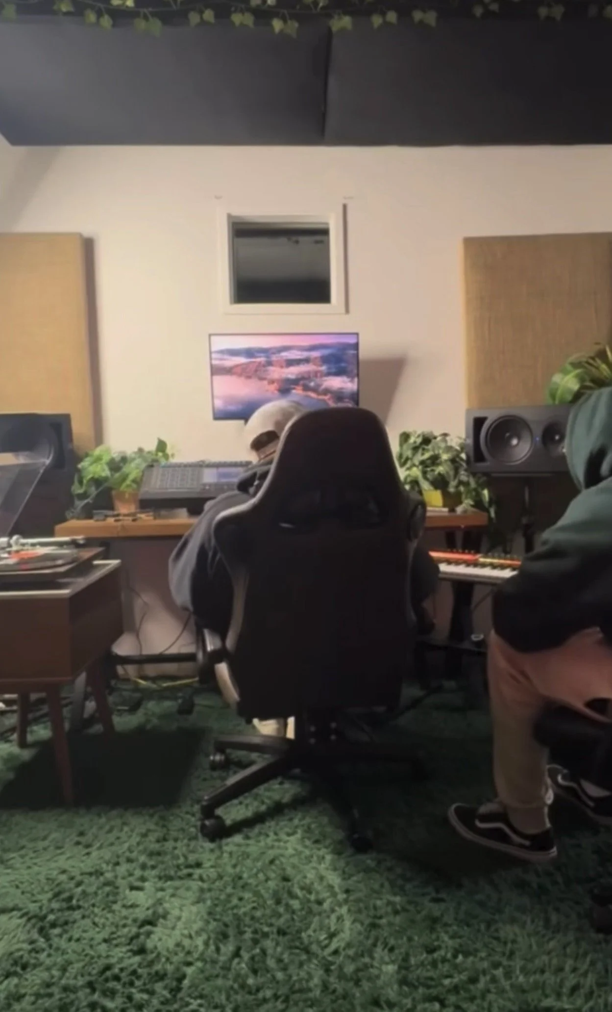 People in a recording studio with soundproof walls, music equipment, and a television displaying a scenic landscape. The room has plants and a green carpet.