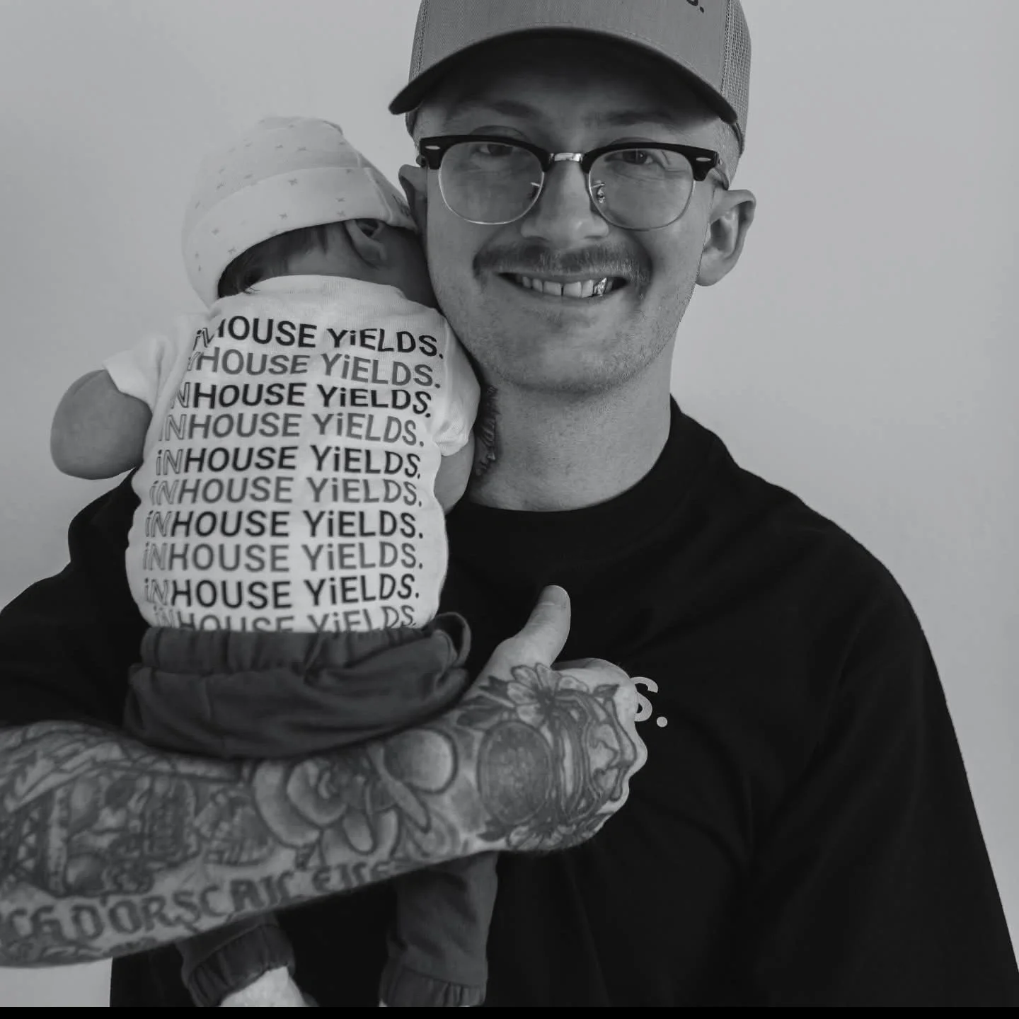 A man wearing glasses and a baseball cap is holding a baby. The baby is wearing a hat and a onesie with the repeated text 'HOUSE YIELDS.' The man has visible tattoos on his arm.