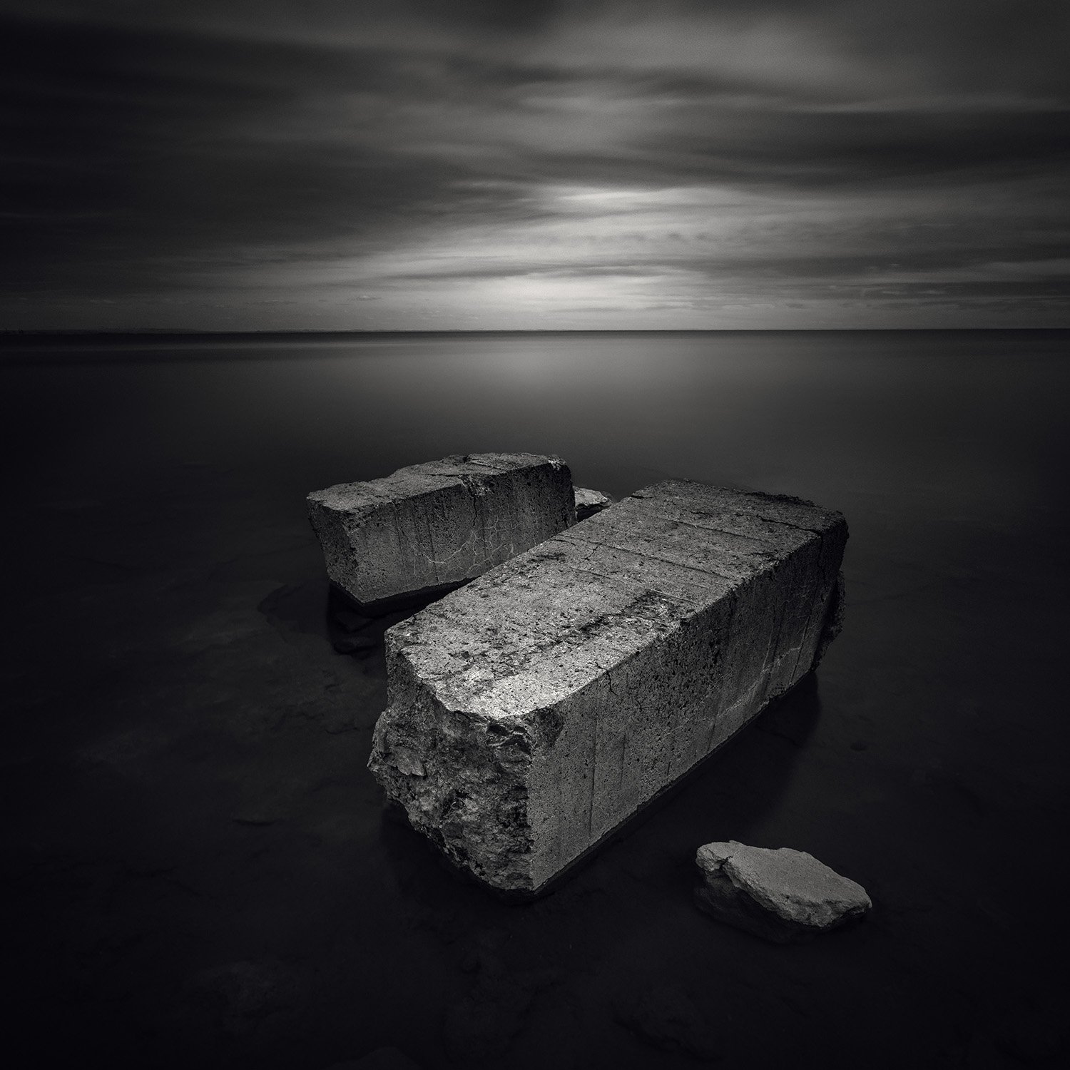 Four large concrete blocks, with one broken, resting on a calm body of water under a cloudy, overcast sky.