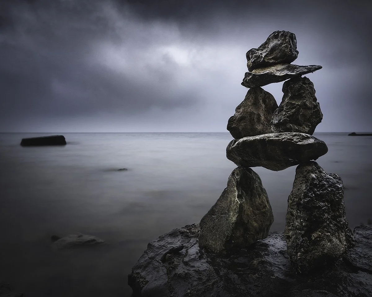 Stacked rocks on a dark, cloudy beach with a calm, misty ocean in the background.