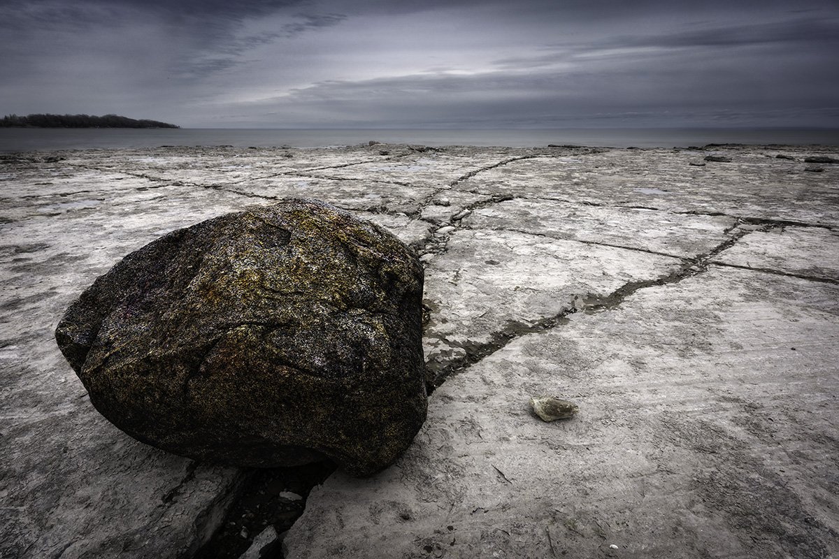 A large, rounded boulder on a flat, cracked, rocky surface near a body of water under a cloudy sky.