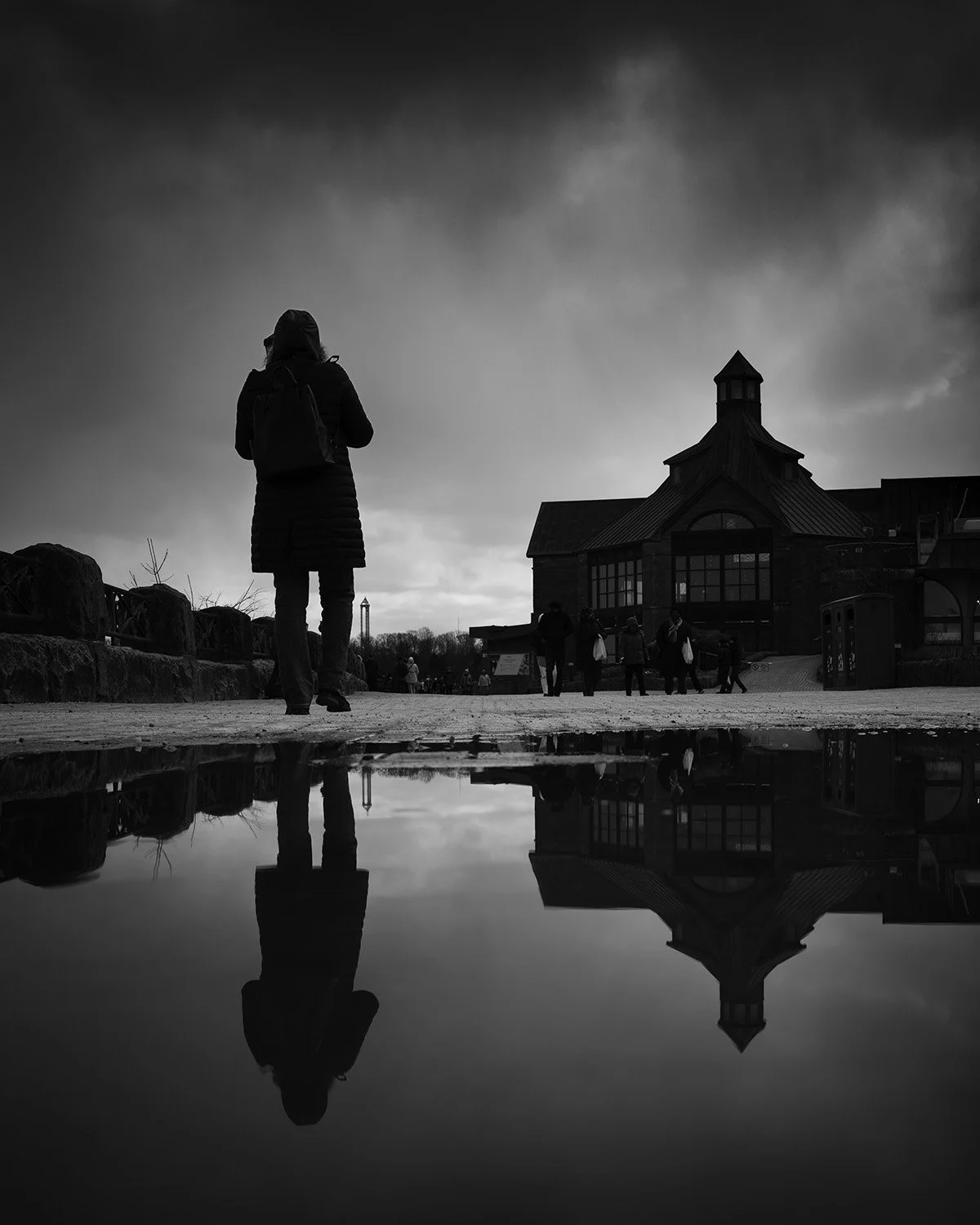 A person wearing a coat and backpack walking towards a building with a peaked roof and lantern tower, reflected in a puddle, with gloomy sky and other people nearby in black and white.