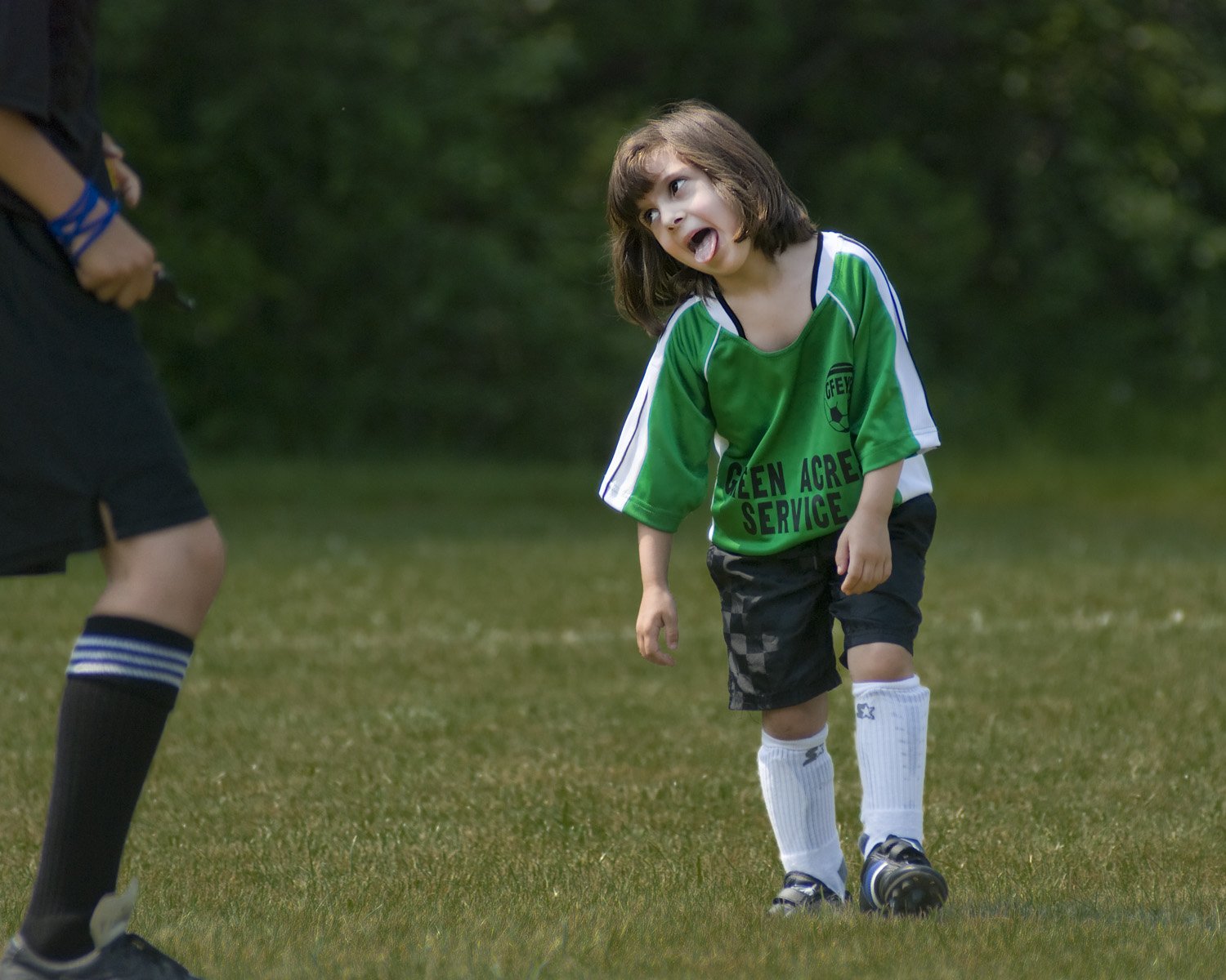 A young girl with shoulder-length brown hair wearing a green soccer jersey, black shorts, white socks, and sneakers standing on a grassy field, leaning slightly forward, appearing tired or upset, with a blurred background of trees and grass.
