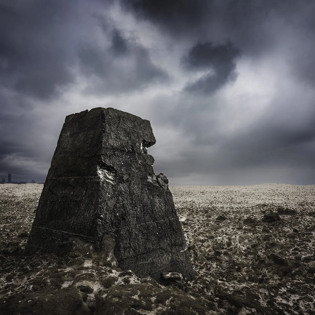 Large weathered rock formation in a barren, rocky landscape with dark, stormy clouds overhead.