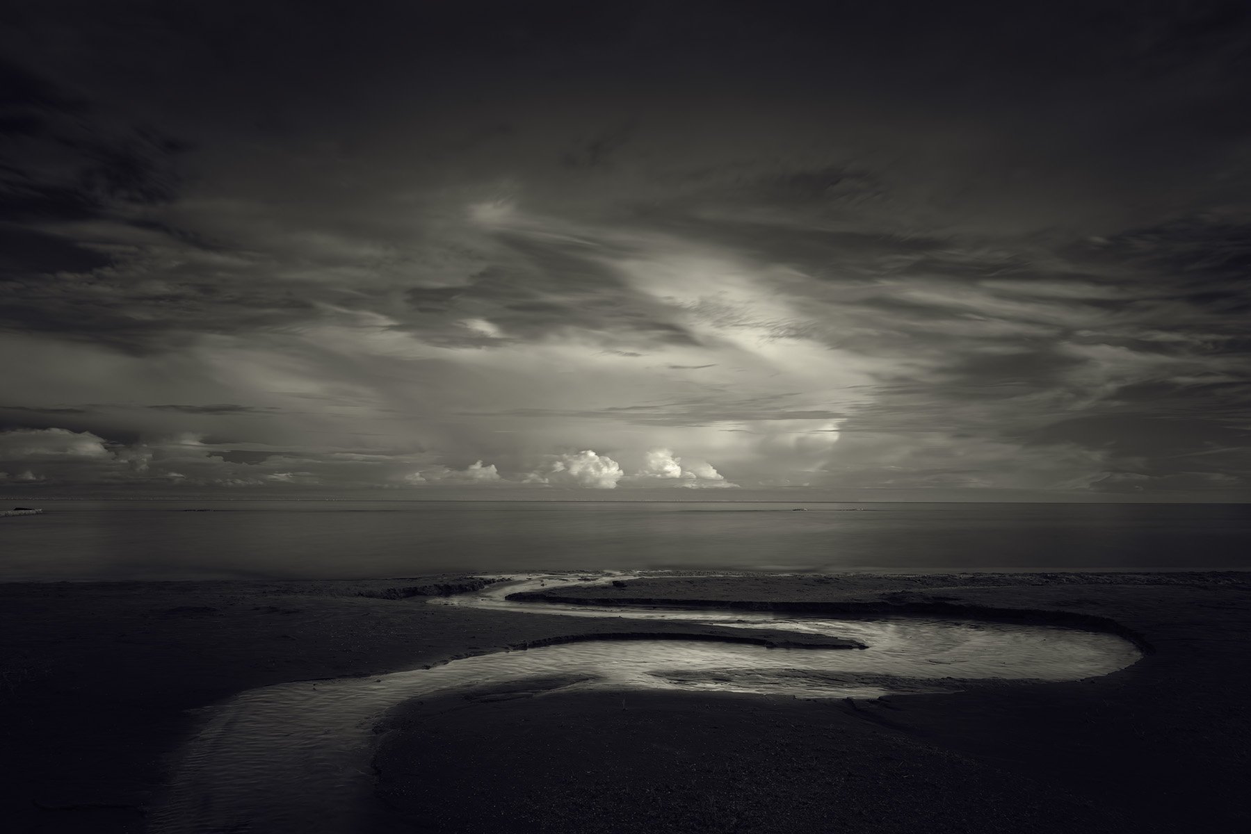 A black and white photo of a beach with a winding river flowing into the ocean under a cloudy sky.