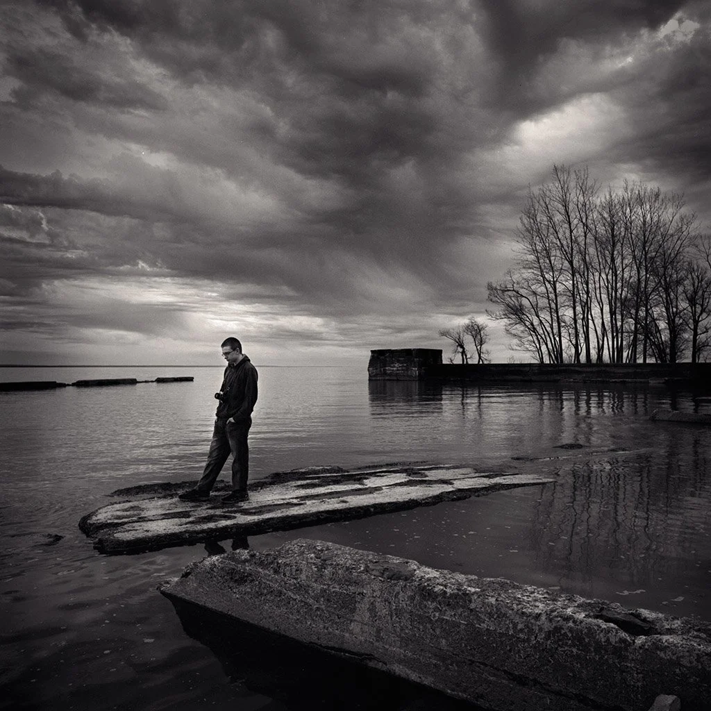 A man stands on an old, weathered wooden dock at the edge of a body of water, looking down or at his phone, with a dark, overcast sky and leafless trees in the background.