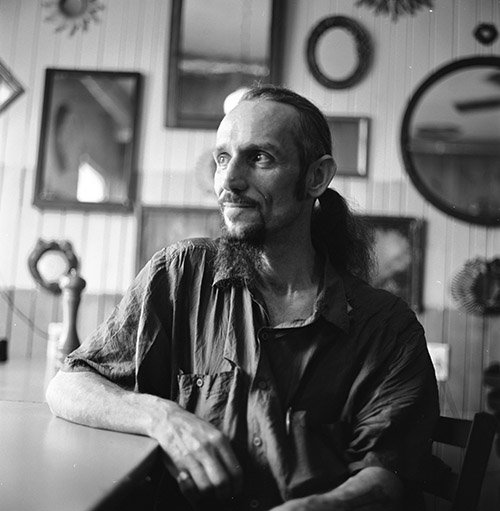 Black and white photo of a man with long hair and a goatee sitting at a table, looking to the side. The background features various framed pictures and decorative items on the wall.