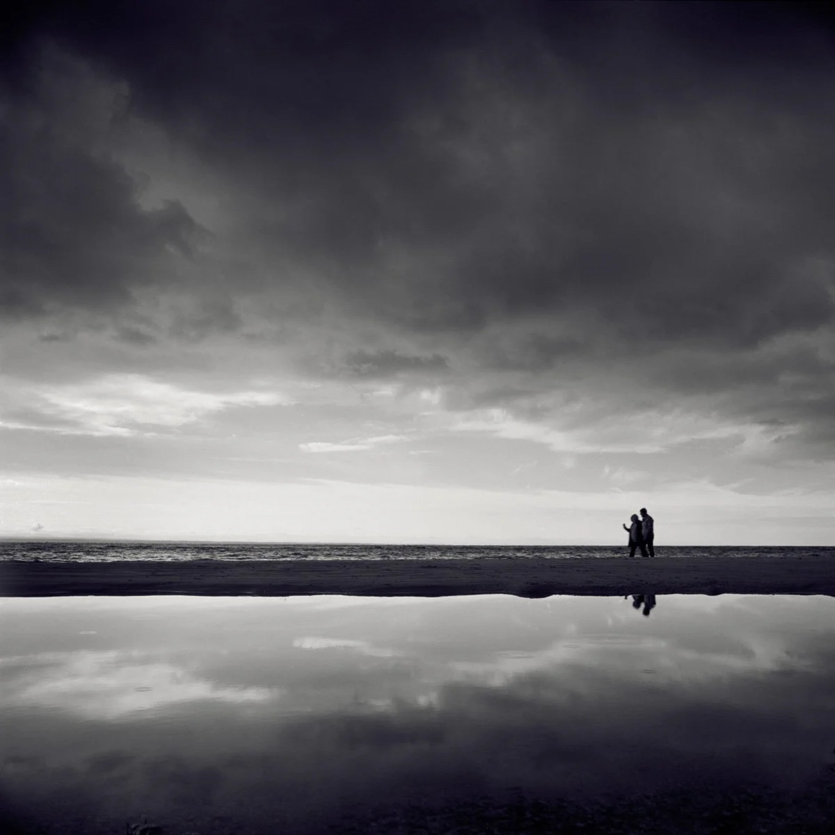 A black and white photo of two people walking along a beach under a cloudy sky, with their reflection visible in a body of water in the foreground.
