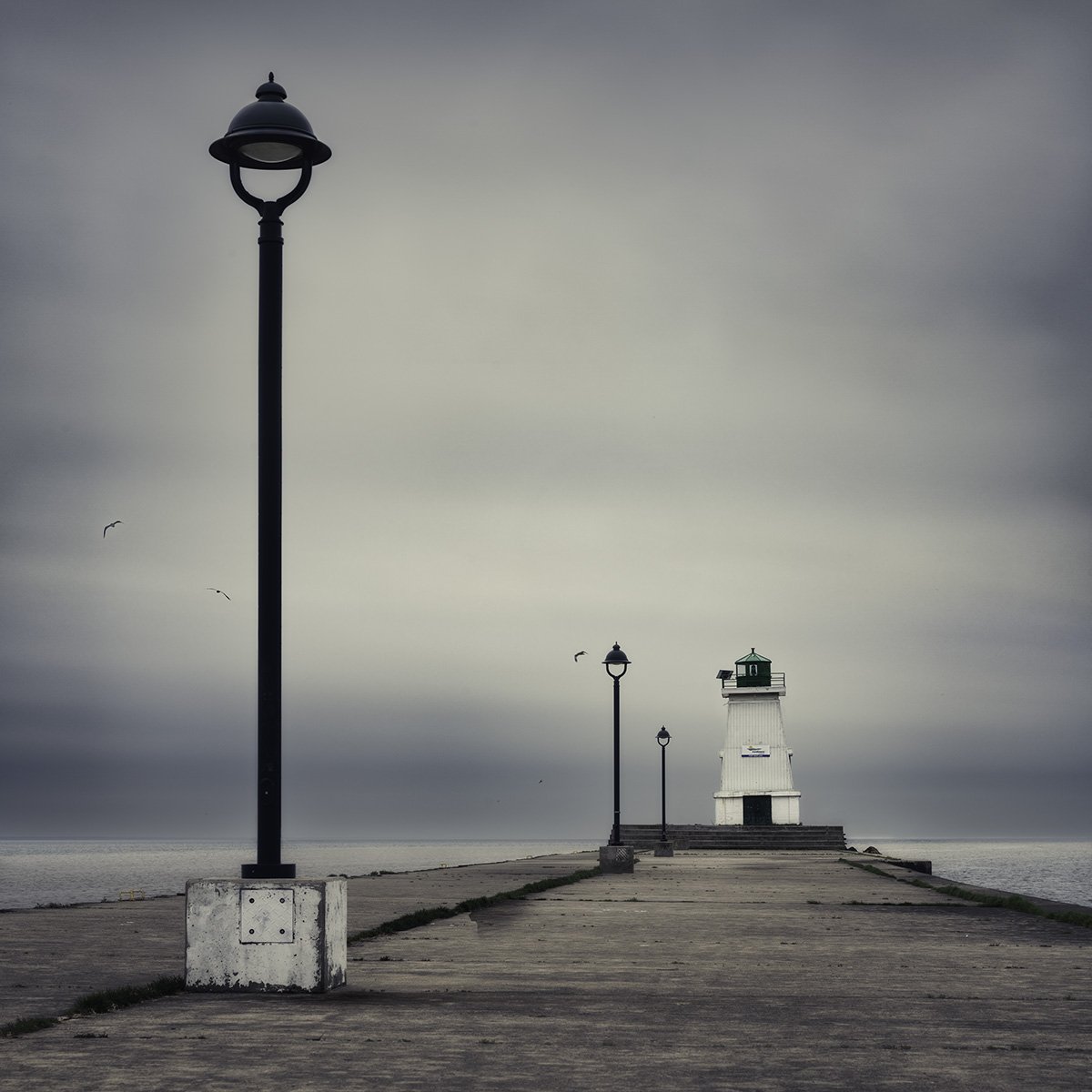 A lighthouse at the end of a pier, with street lamps along the walkway, under a cloudy sky, with seagulls flying nearby.