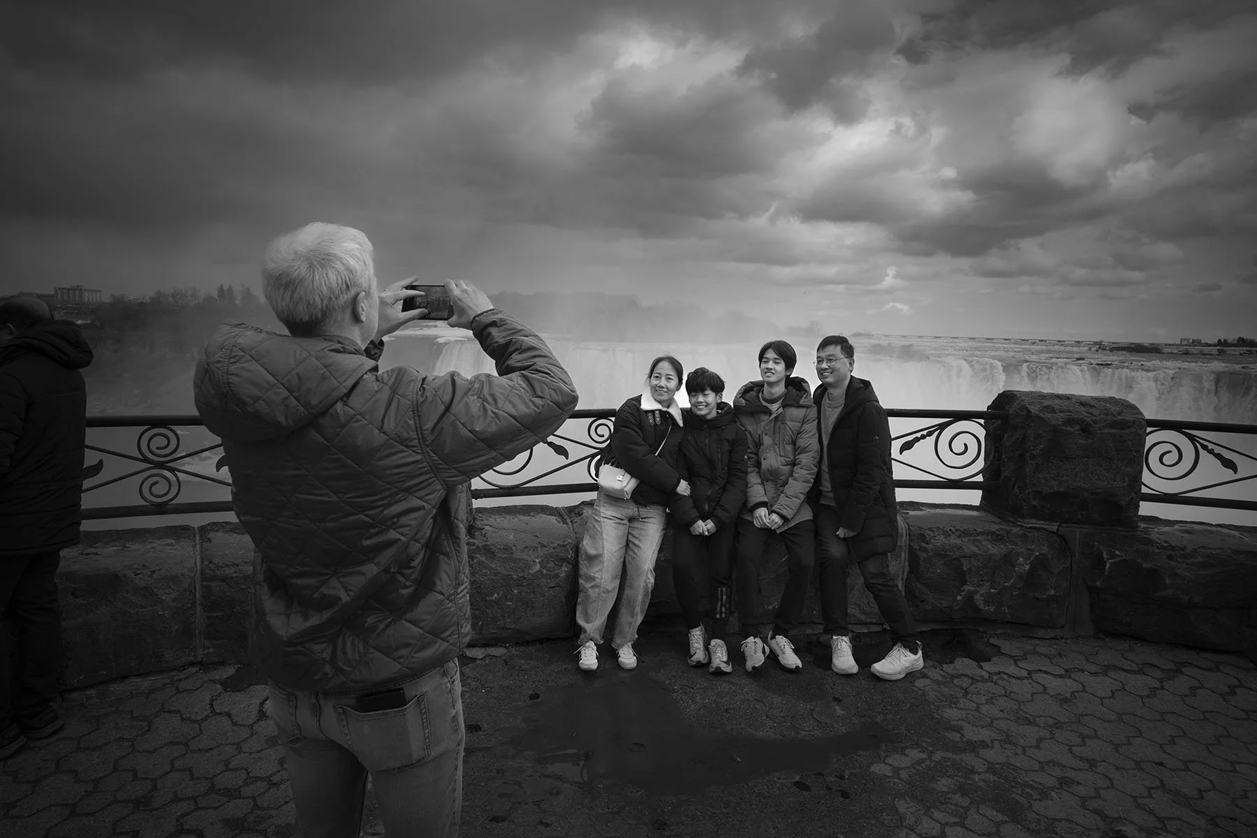 A group of five people posing for a photo at Niagara Falls, with a man taking the picture. The scene features a cloudy sky and a railing, and the people are smiling and dressed in warm clothing.