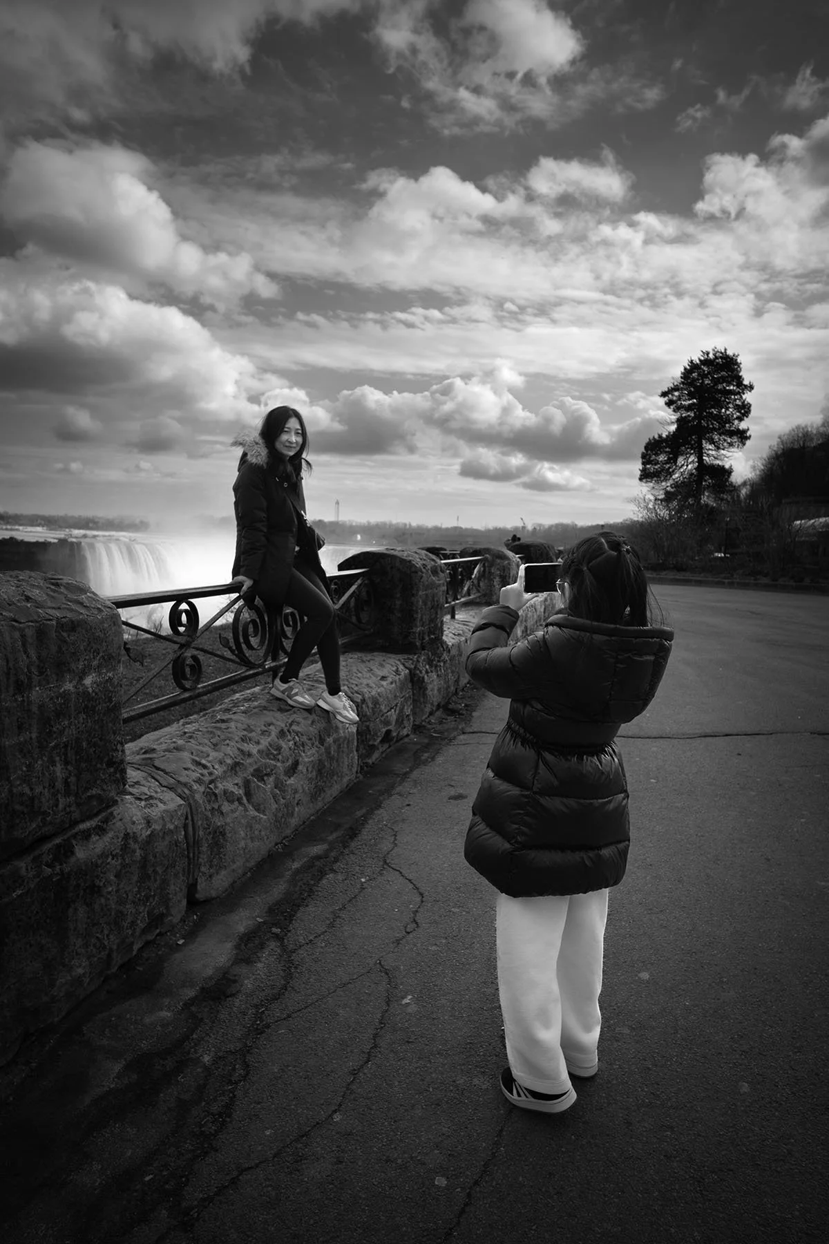 A woman leaning on a stone and metal railing by a waterfall, being photographed by another person on a cloudy day.