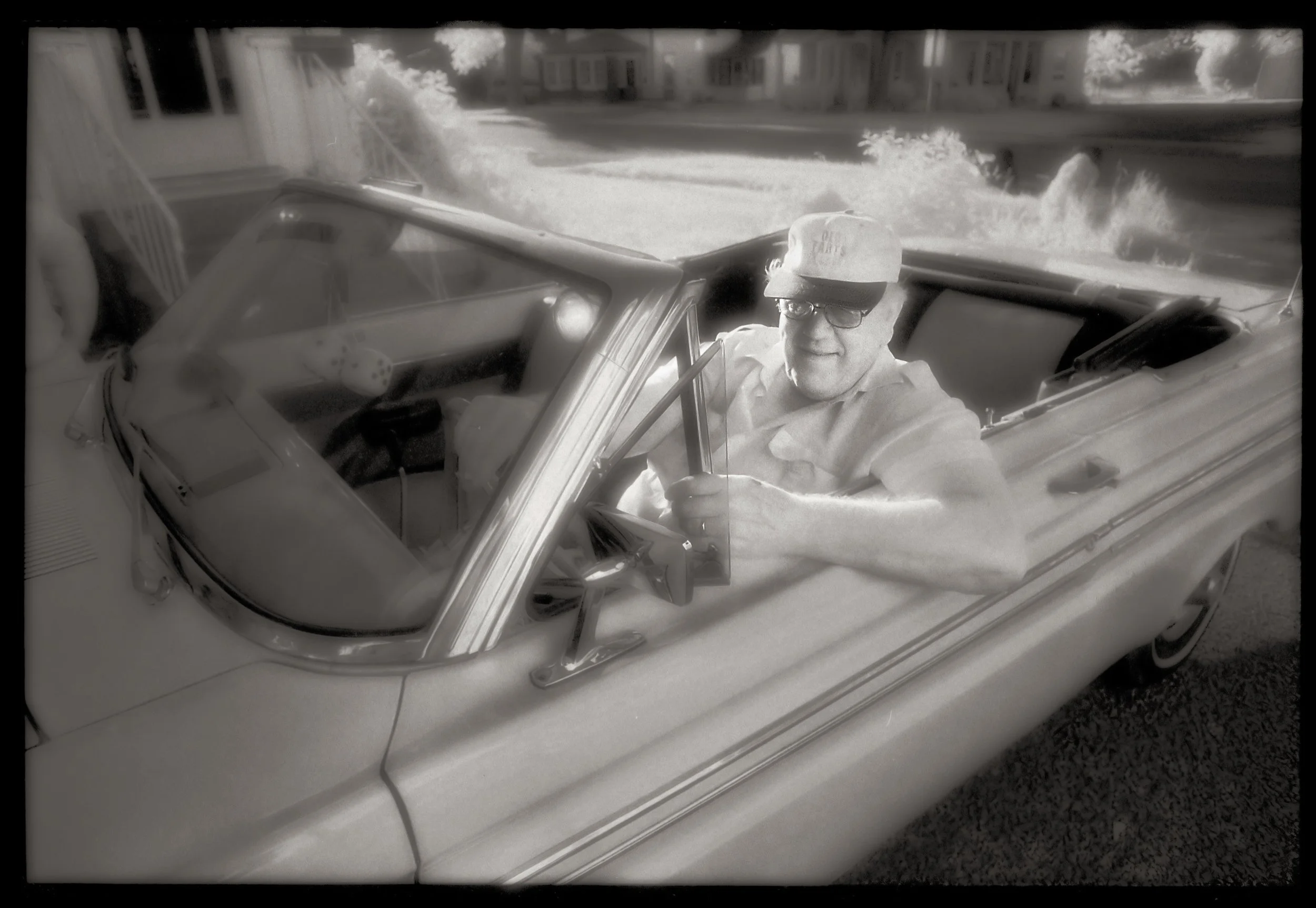 A black-and-white photo of a man sitting in a vintage convertible car, wearing glasses, a cap, and a light-colored shirt, with a residential neighborhood in the background.