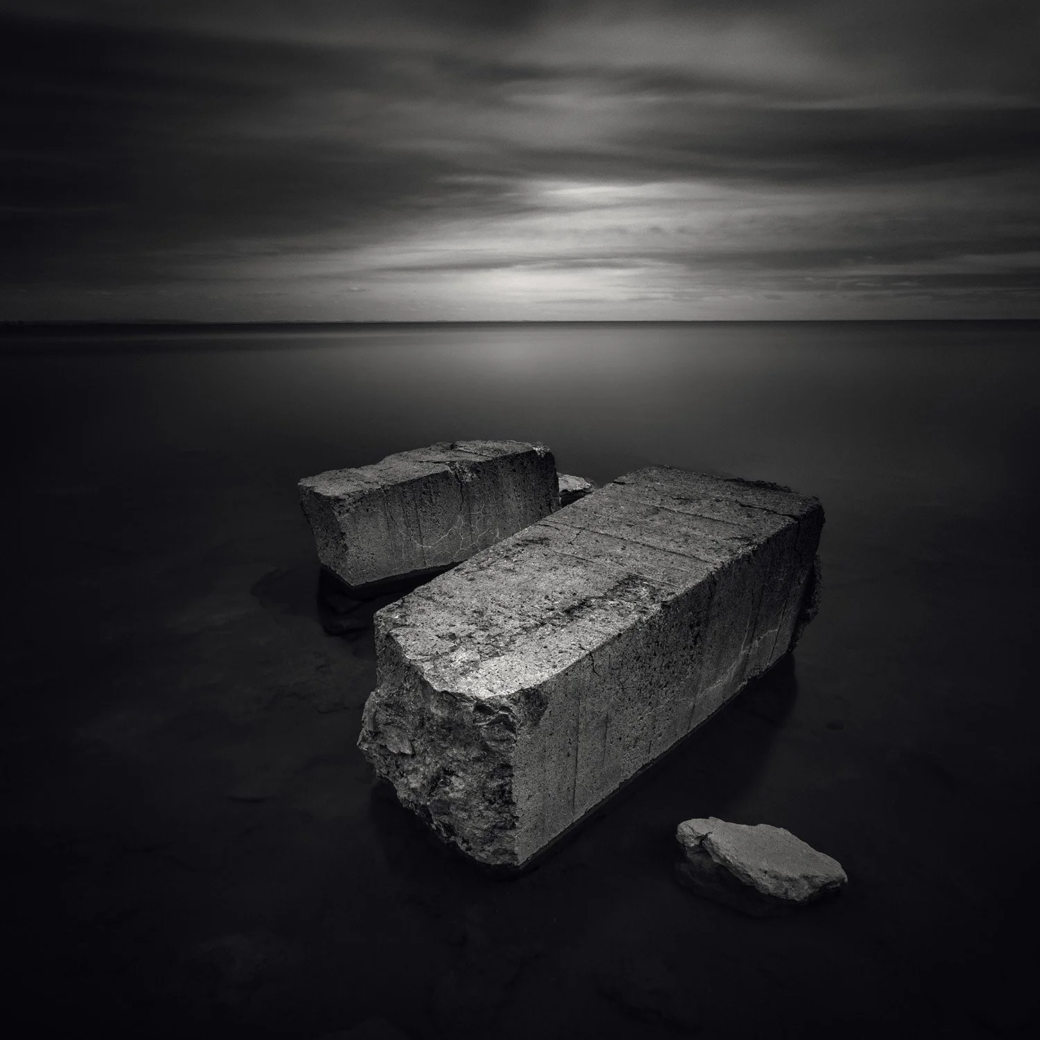 Three large concrete blocks on a dark shore with calm water and a cloudy sky in the background.