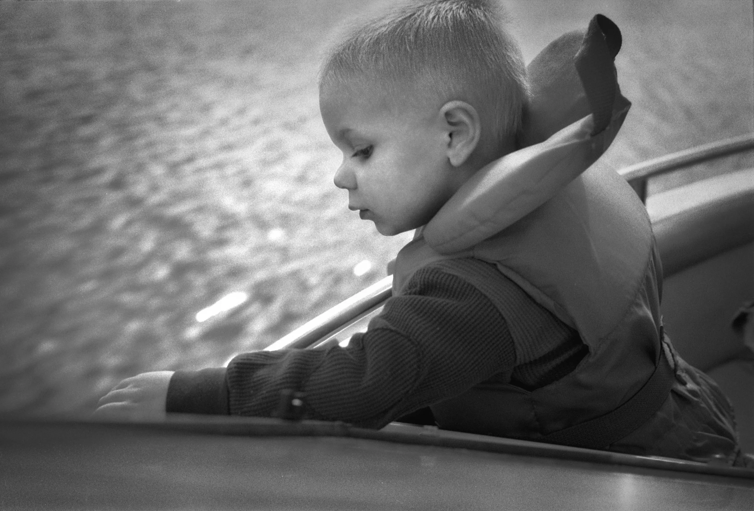 A young boy with short hair leaning out of a car window, looking down, with water in the background.