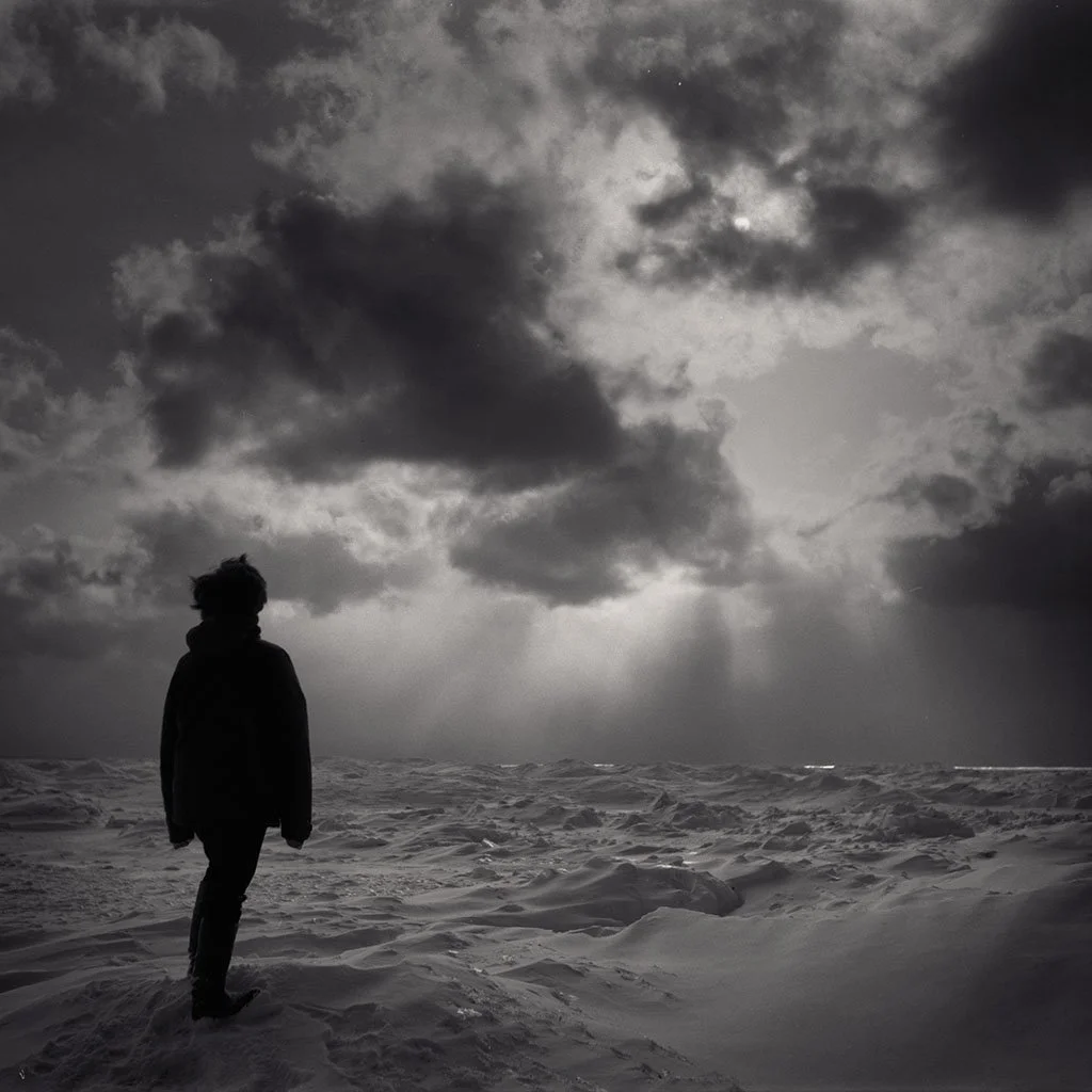 A person standing on snowy terrain under a cloudy sky with rays of light breaking through the clouds.