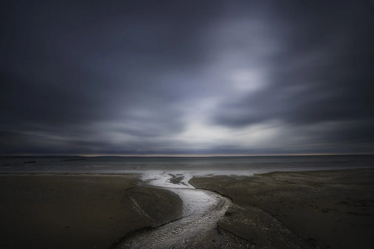 A dark, cloudy sky over a beach with a small stream of water flowing toward the ocean.