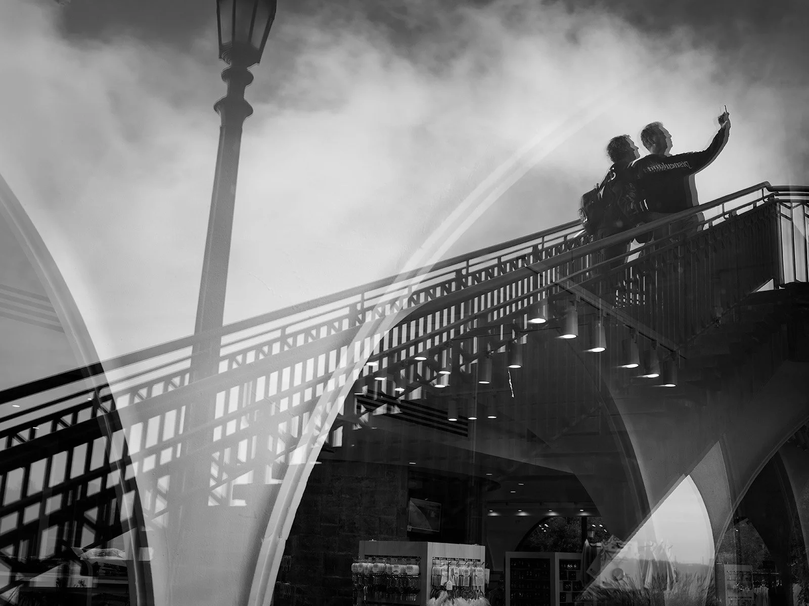 Two people taking a selfie or photo together on an elevated walkway with street lighting and cloudy sky visible in the background.