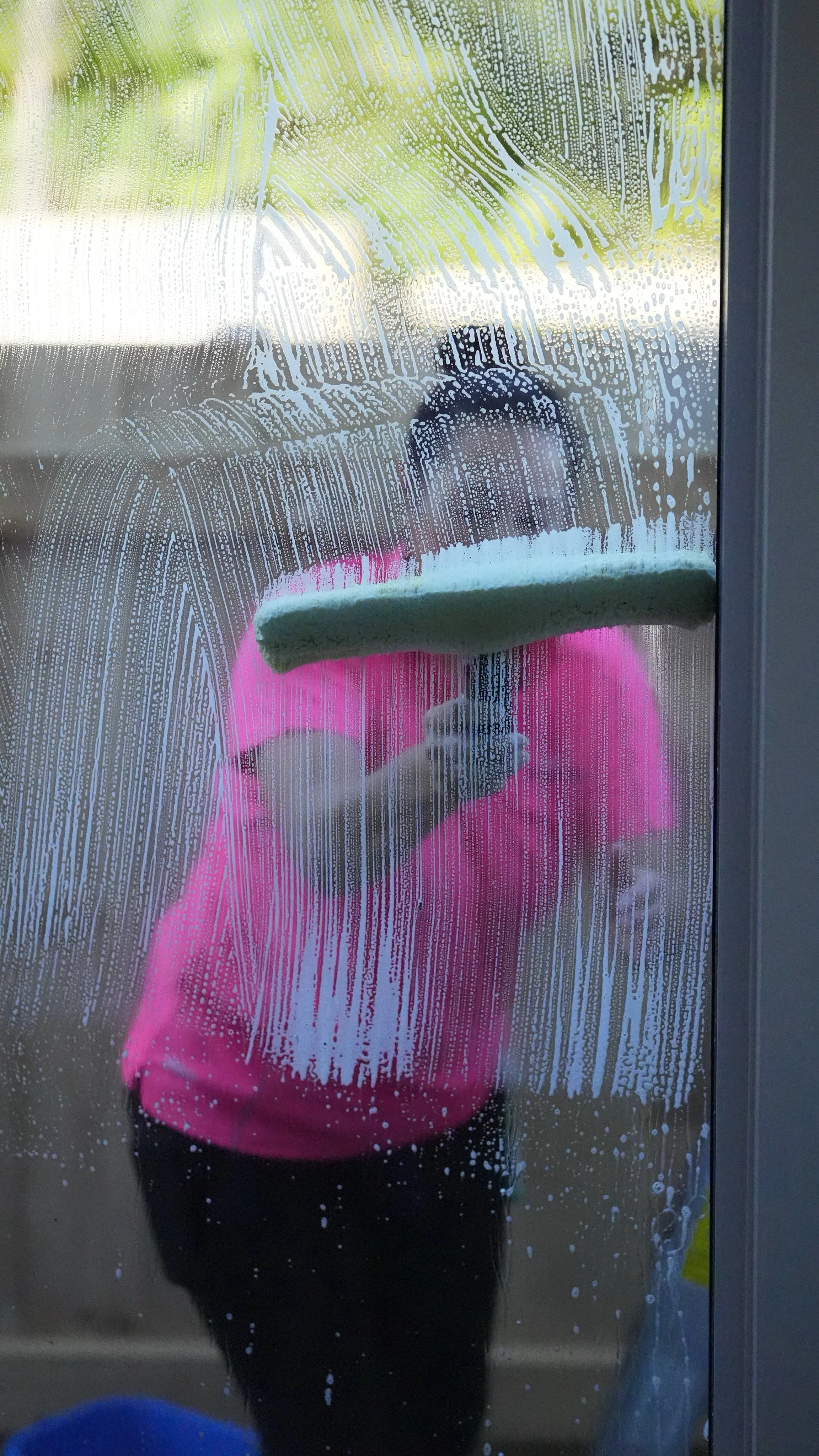 Person wearing a pink shirt is cleaning a glass door with a yellow sponge, with soap and water. Reflections of the person and a green outdoor scene are visible on the glass.
