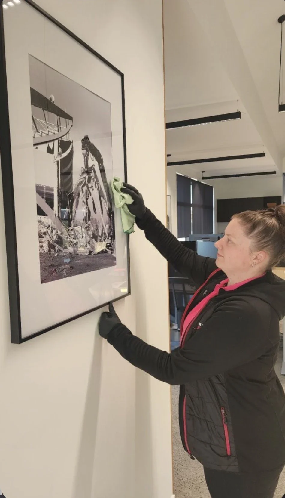 A woman wearing black gloves cleaning a framed black and white photograph on a wall in a gallery or office space.