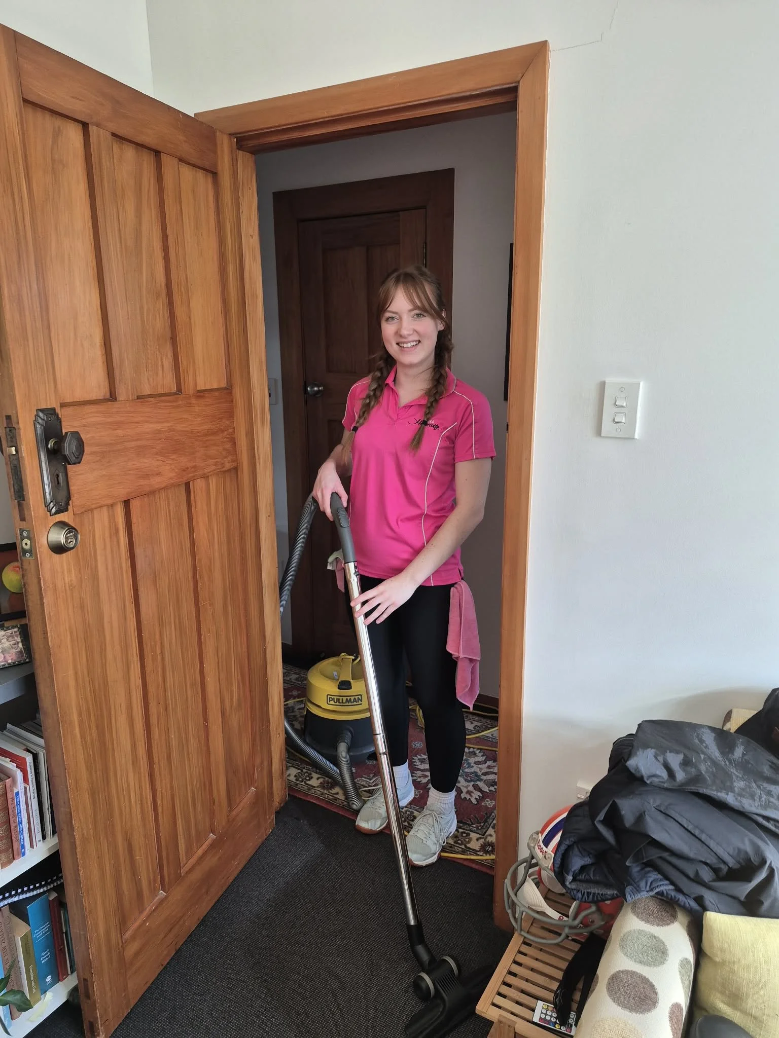 A young woman with braided hair wearing a pink shirt vacuuming a room with a yellow vacuum cleaner. She is smiling and standing in the doorway of a room with wooden door frames and a patterned rug.
