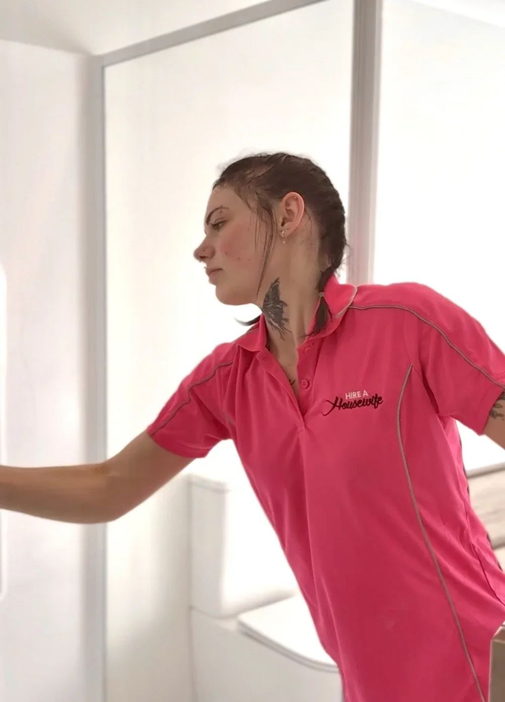 A woman wearing a pink shirt with the text "Hire a Houswife" on it, reaching for something in a room with white walls and a toilet in the background.