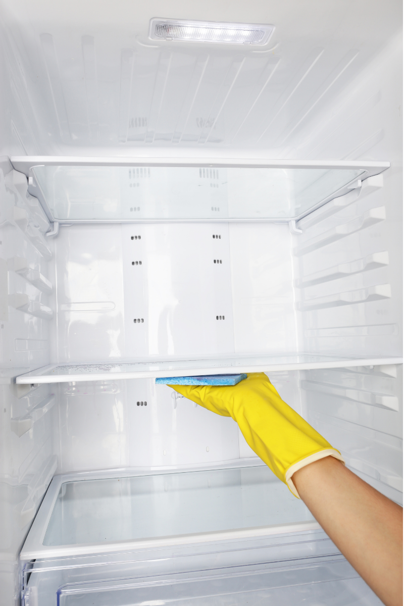 A person wearing a yellow rubber glove cleaning the inside of a refrigerator with a blue sponge.