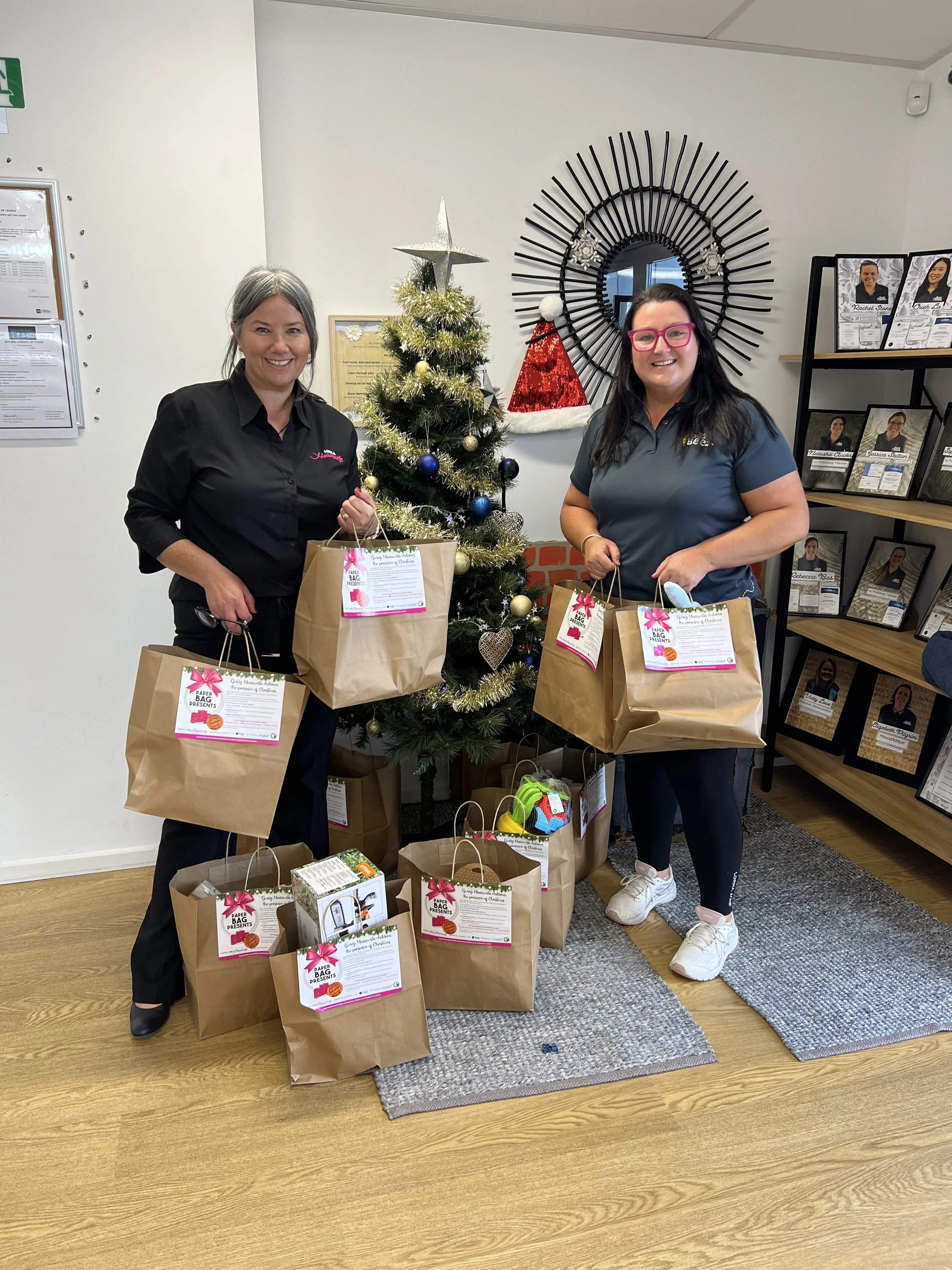 Two women standing next to a Christmas tree holding paper gift bags, with more gift bags on the floor in front of them. The woman on the left has gray hair and black hair, and the woman on the right has black hair and pink glasses. Behind them, a wall features a decorative black circular mirror, holiday decorations, and framed photos of people.