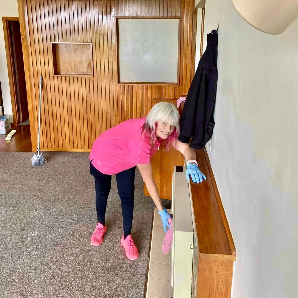 A woman with gray hair, wearing a pink shirt, black pants, pink shoes, and blue gloves, cleaning a wooden ledge in a room with wood-paneled walls and a carpeted floor.