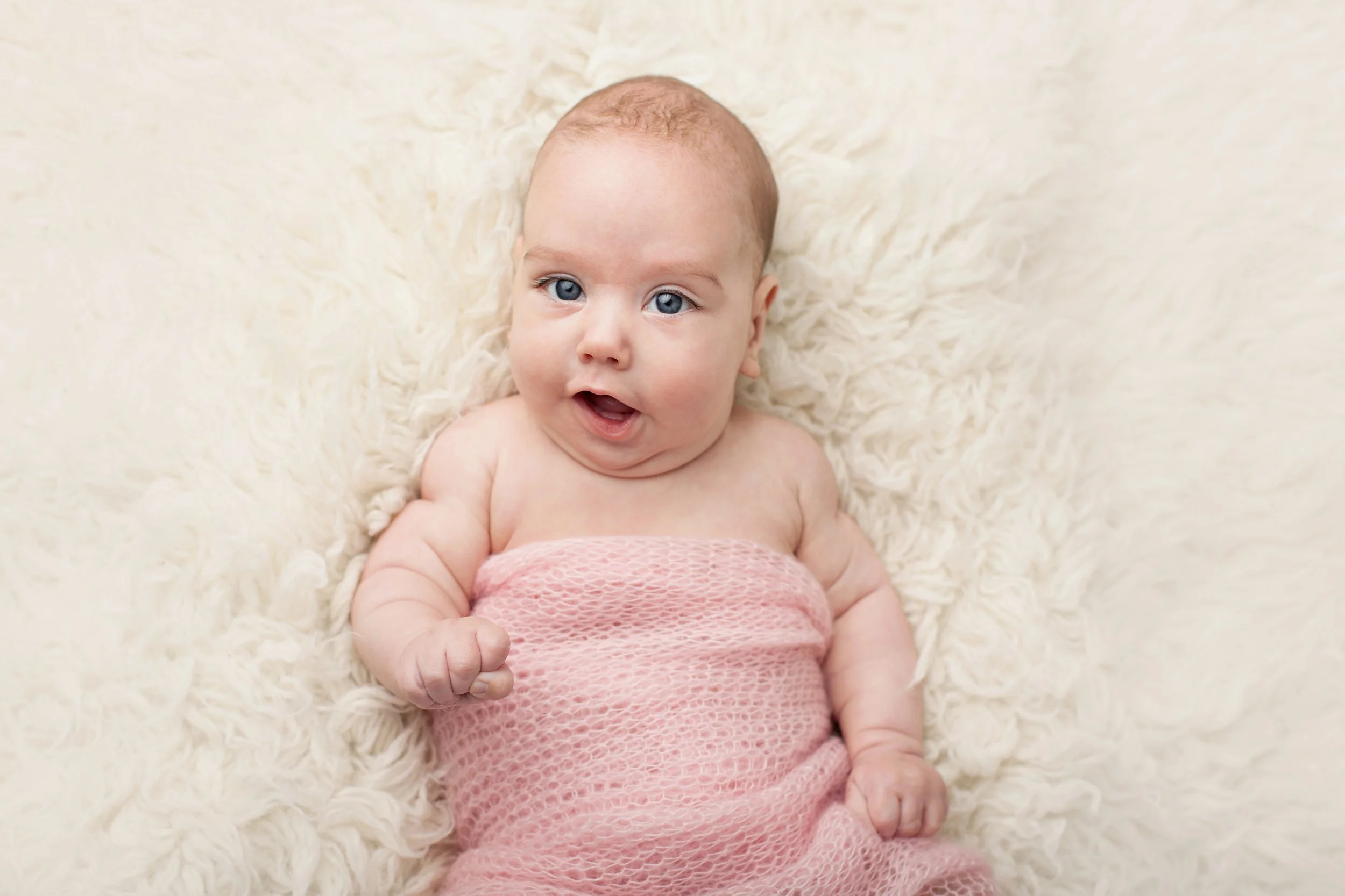 A baby with blue eyes and light skin lying on a fluffy cream-colored blanket, wearing a pink textured wrap, looking at the camera with an open mouth.
