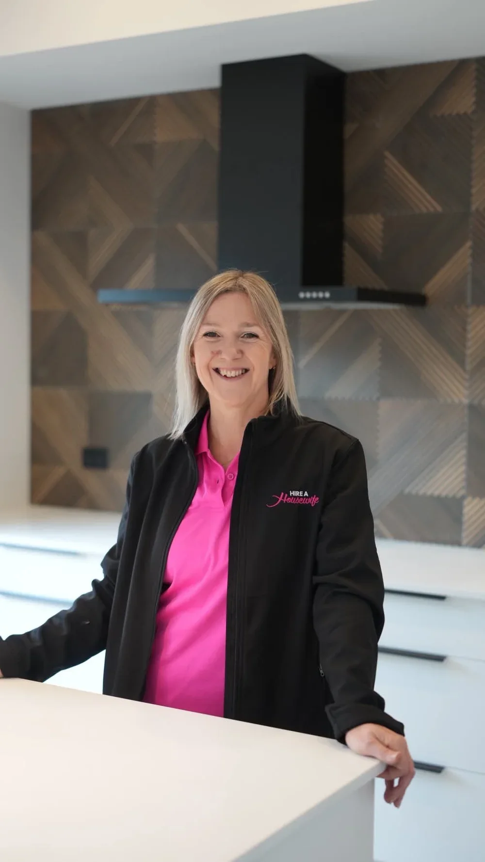 A smiling woman in a pink shirt and black jacket with a logo that reads 'Hire a Housewife' stands behind a white counter in a modern kitchen with dark wooden herringbone-patterned backsplash.