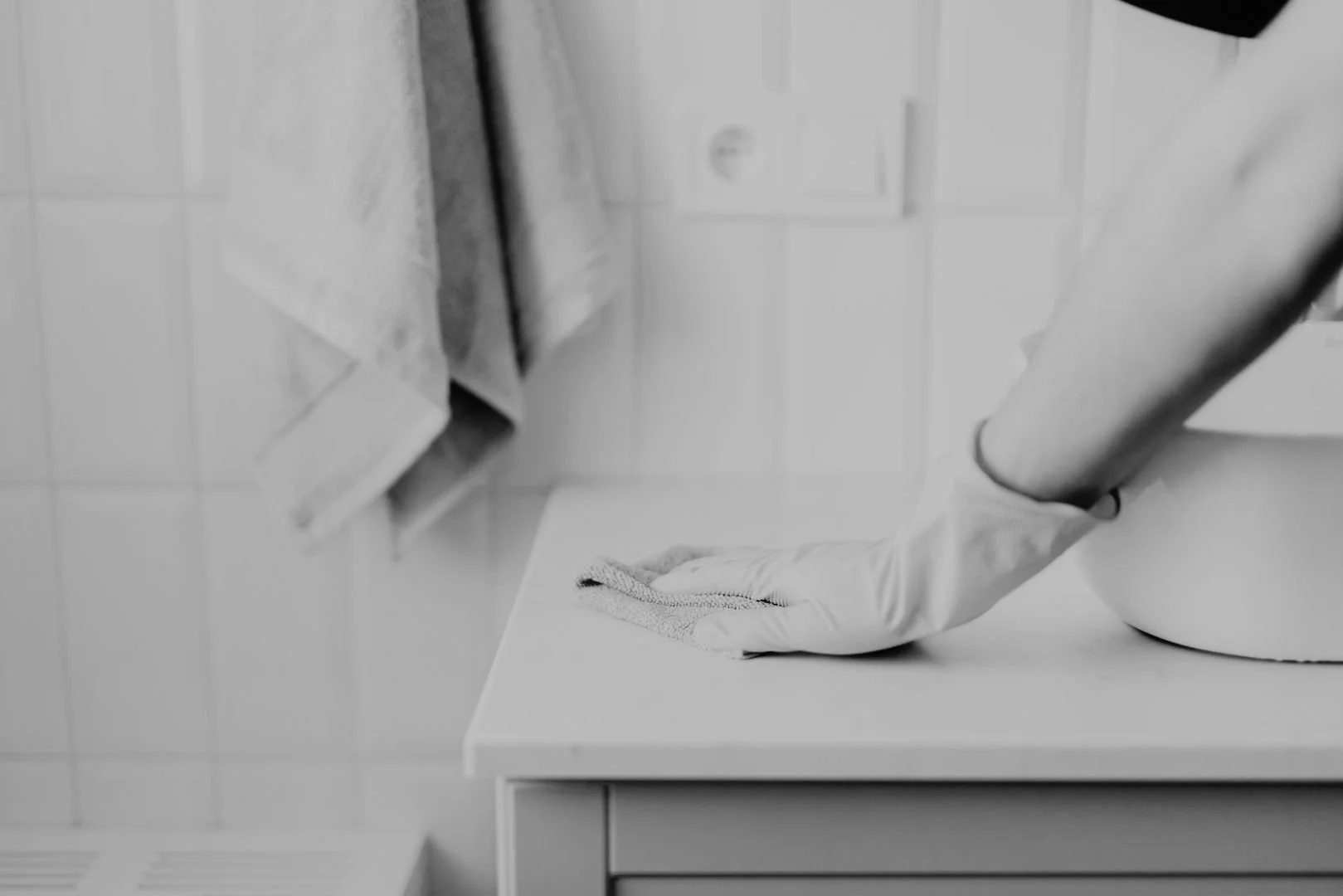 Person wearing gloves cleaning a white surface with a cloth in a bathroom or kitchen setting.