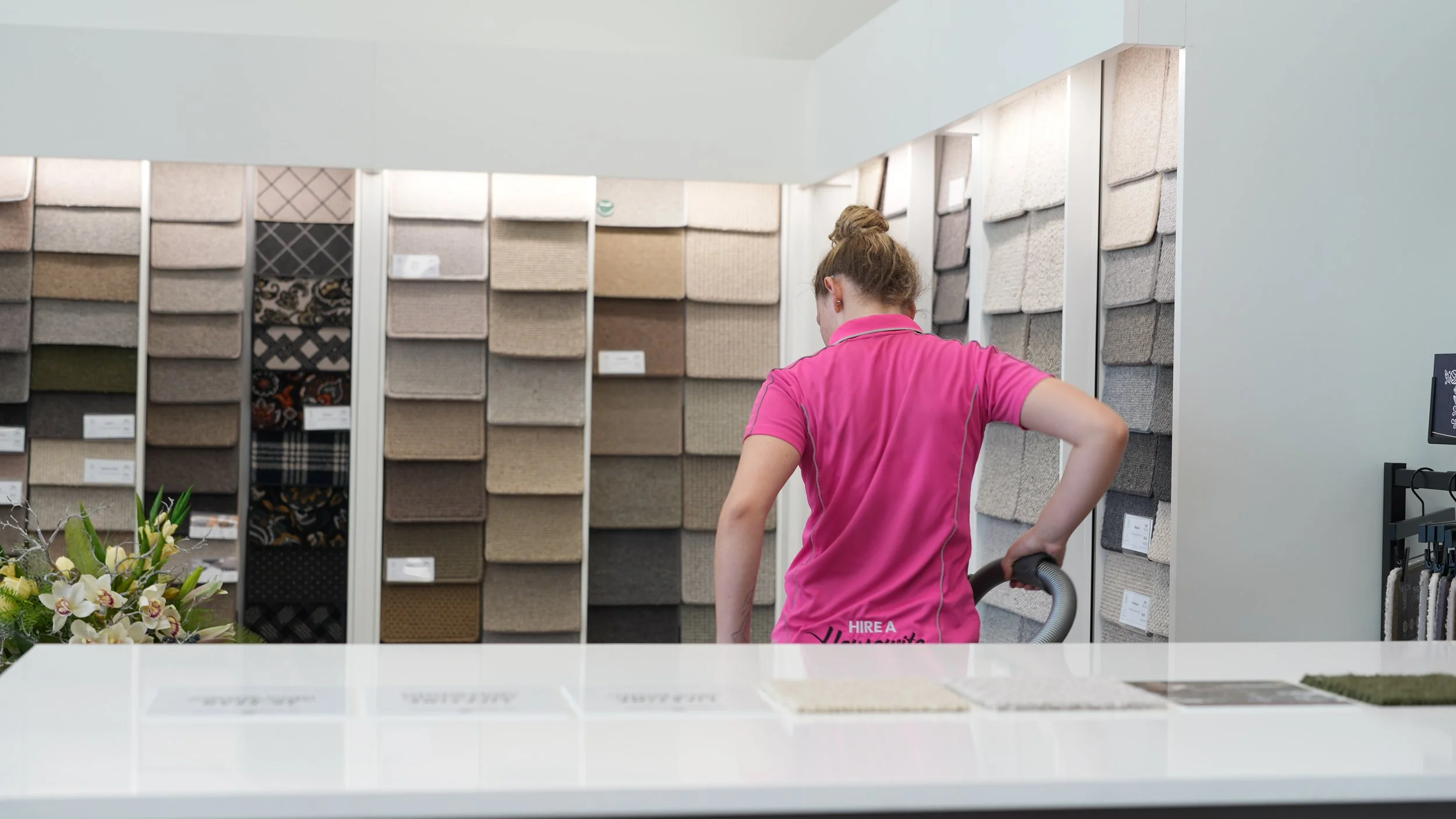 A woman in a pink shirt vacuuming in a carpet store with fabric samples on display behind her.