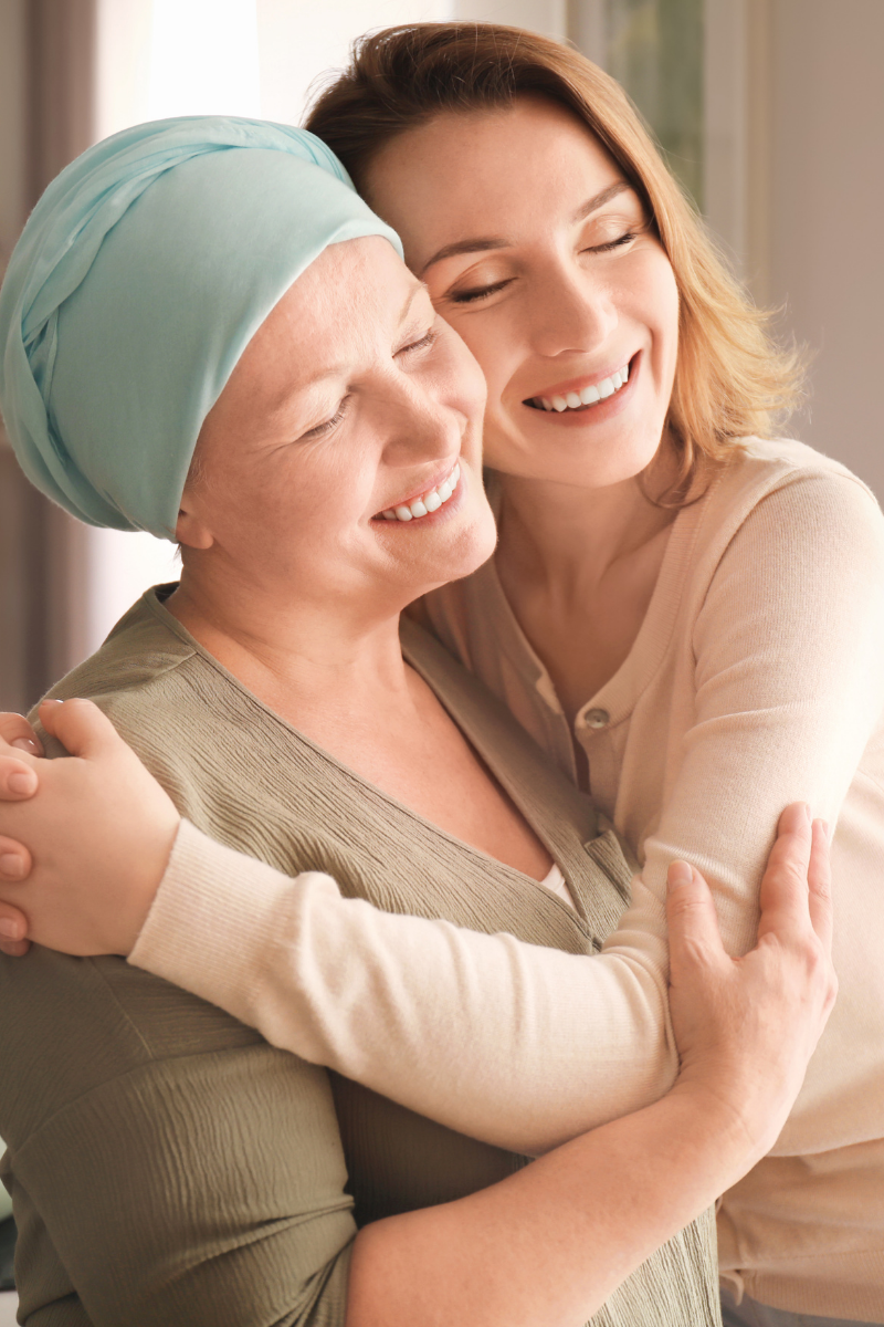 An elderly woman with a turquoise headscarf hugging a young woman with red hair, both smiling and enjoying a loving embrace.
