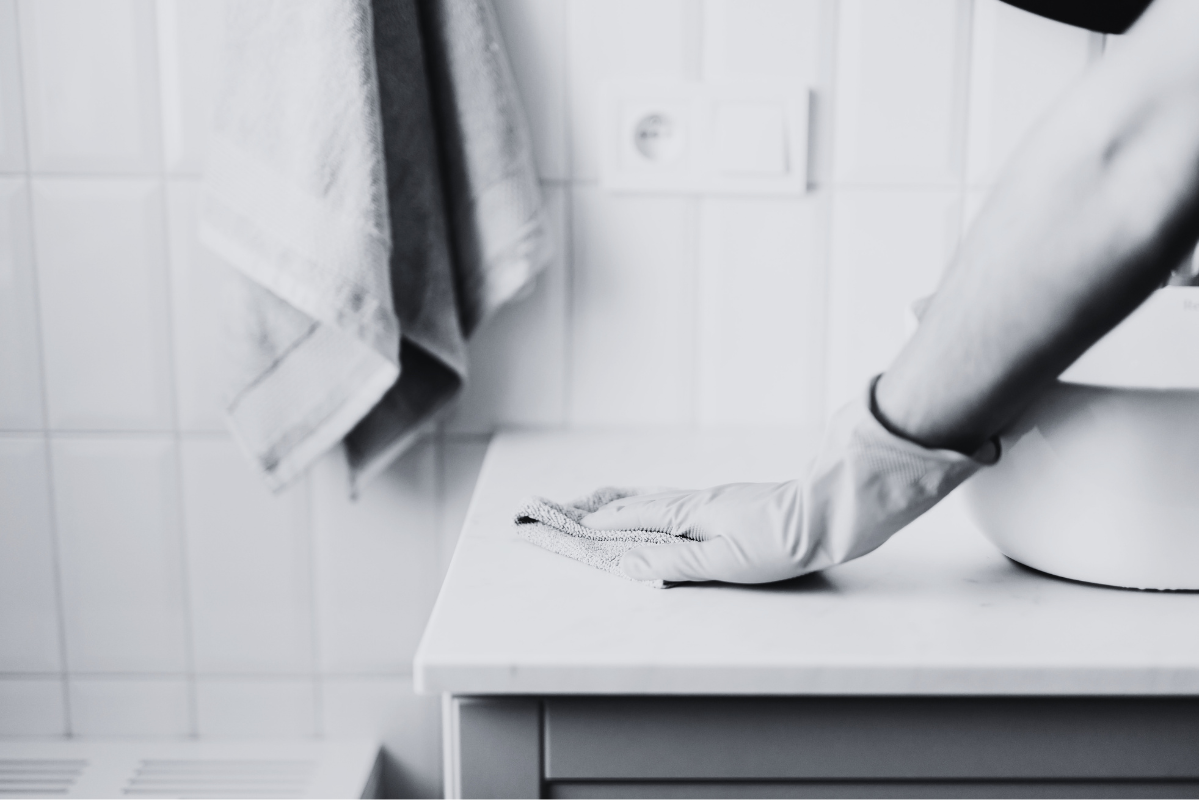 Person wearing gloves cleaning a white surface in a bathroom with towels hanging on the wall.