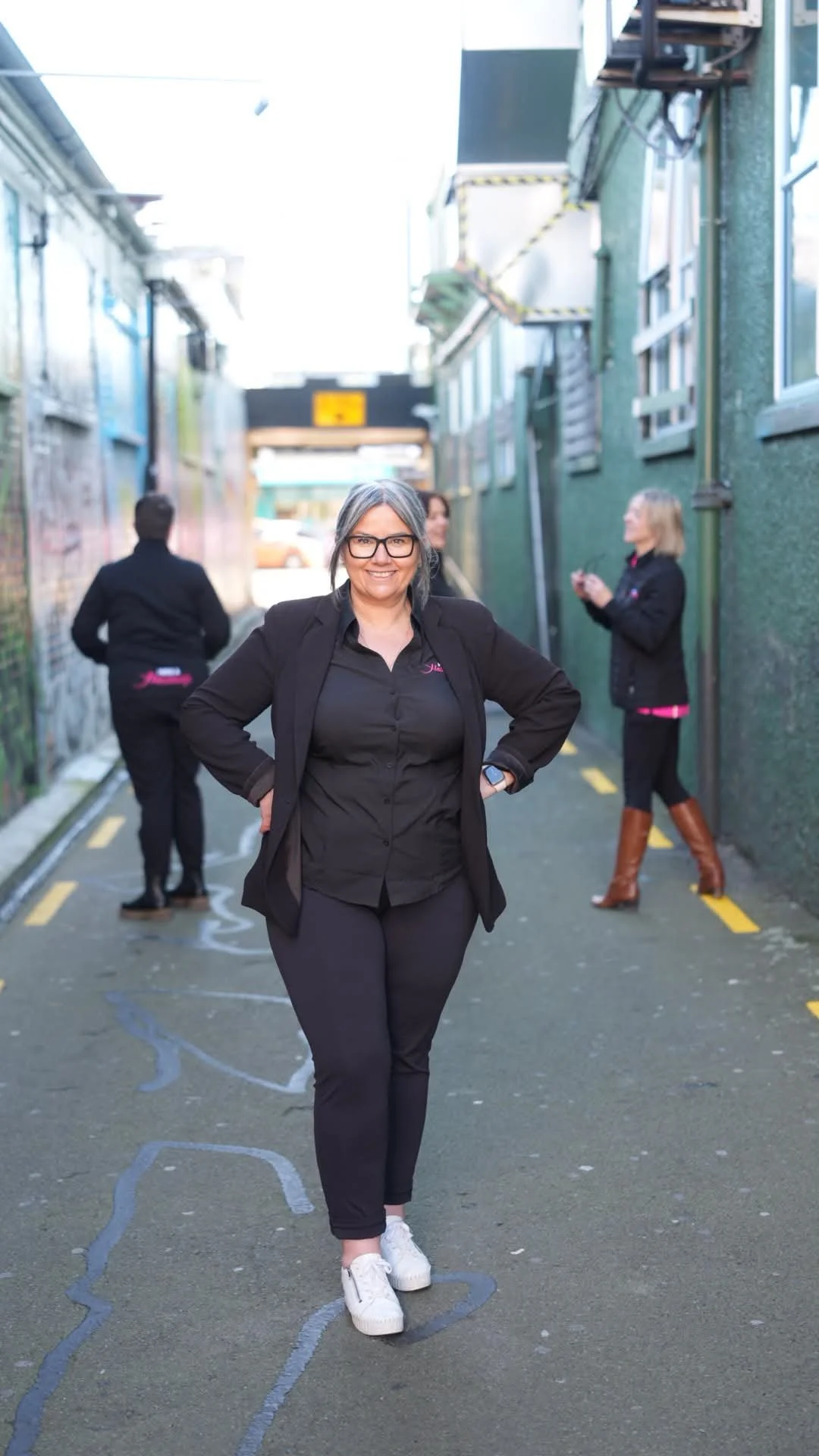 A woman with gray hair, glasses, and a black blazer smiling with hands on hips in an alleyway with graffiti on the walls, with three other women in the background, two of whom are looking at their phones.