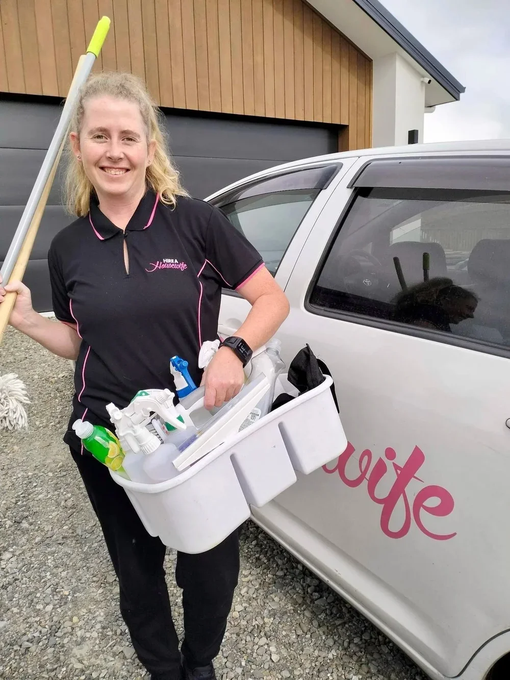 A woman smiling, holding a cleaning spray bottle and a bucket of cleaning supplies, standing next to a white car with the word 'Wife' partially visible on its side, in a driveway in front of a house.