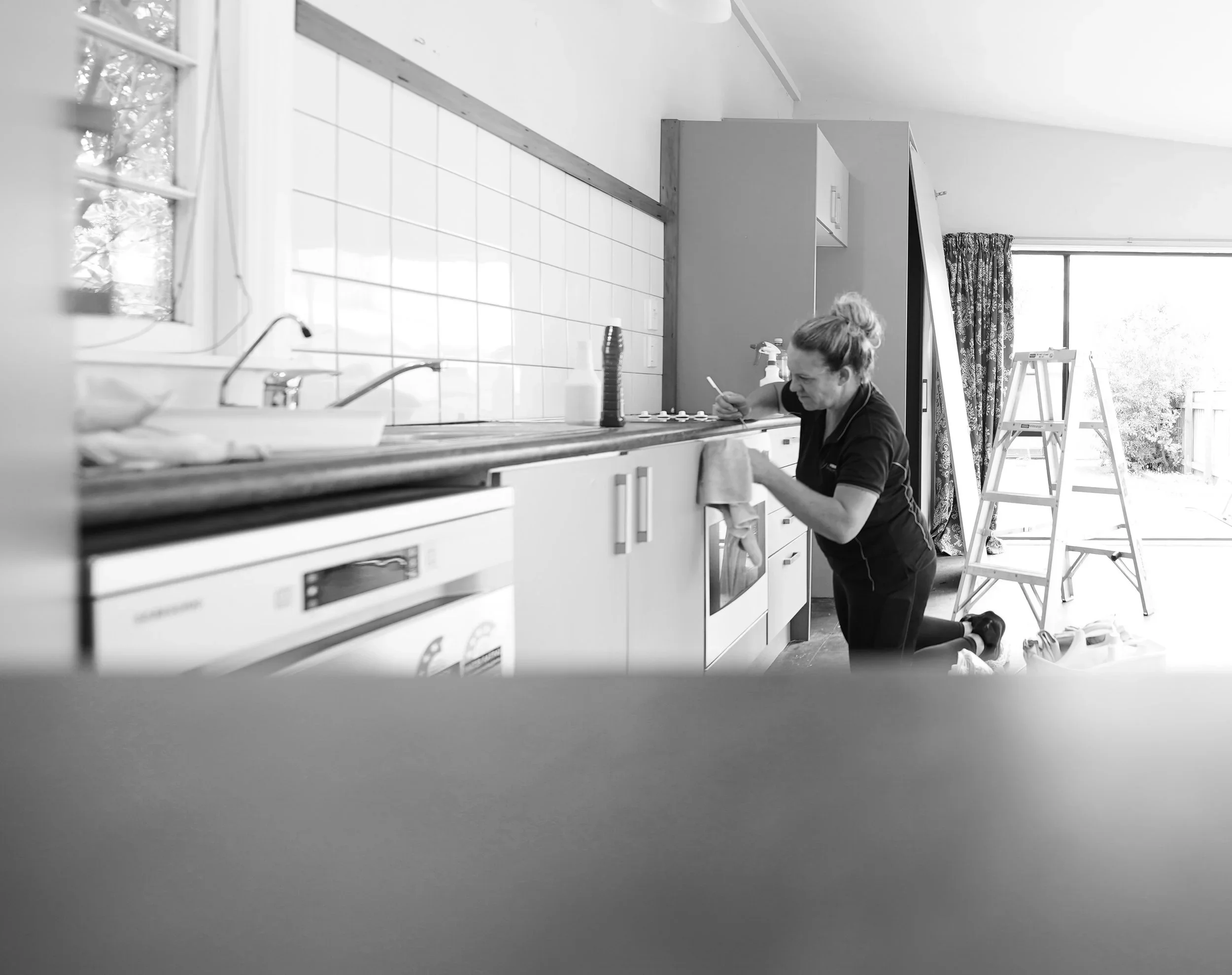 A person kneeling on the floor in a kitchen, writing on a surface, with ladders and a window in the background.