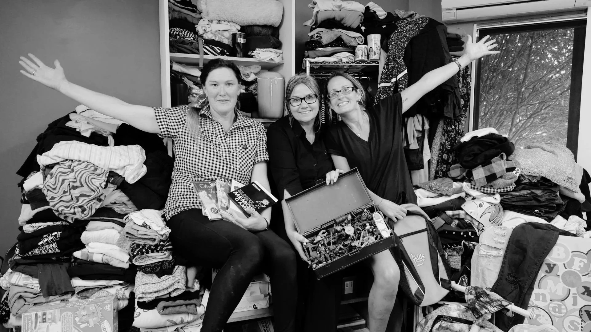 Three women sitting on piles of clothing and toys in a room, with open shelves of laundry and a window showing trees outside. The women are smiling; one is holding a box of building blocks, one is holding a collection of toy cars, and the third is gesturing with arms wide open.