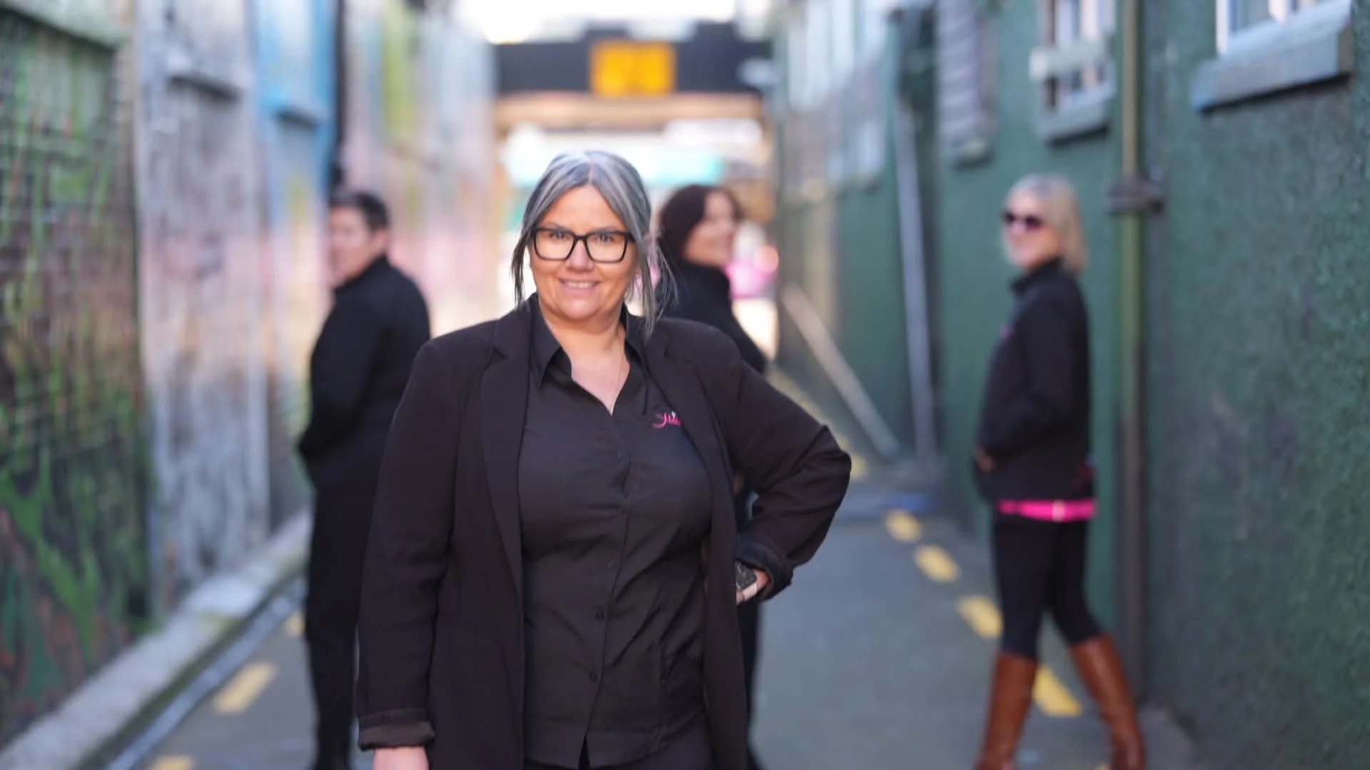 Four women standing in an alleyway. The woman in front is smiling, wearing glasses, a black blazer over a black shirt. The other women are in the background, also dressed in black, with two of them looking to the side and one wearing sunglasses. The alley has a graffiti-covered wall on one side and a green fence on the other, with a yellow line painted on the ground.